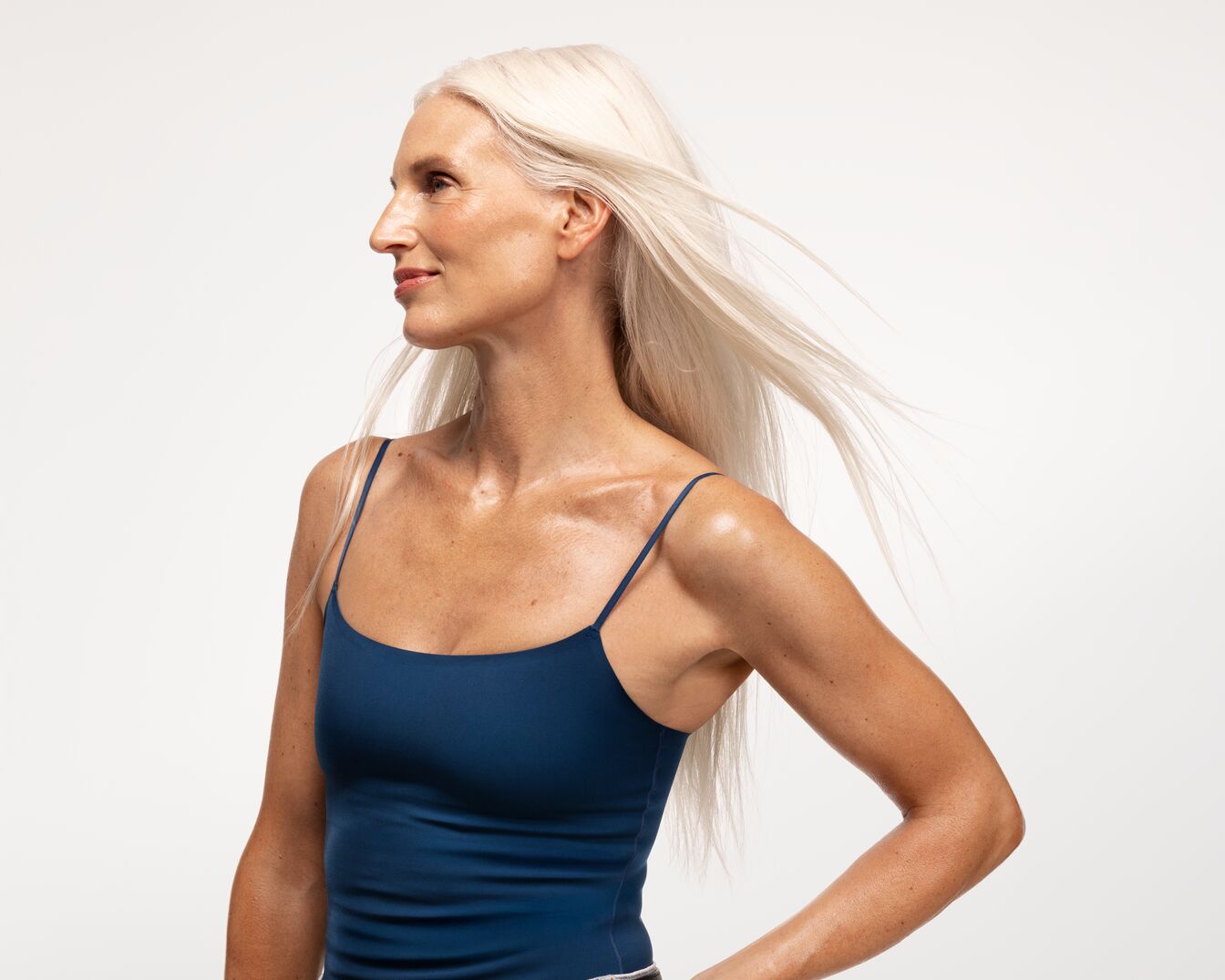 Eine Frau mit langen, weißen Haaren und einem blauen Tanktop posiert vor einem hellen Hintergrund.
