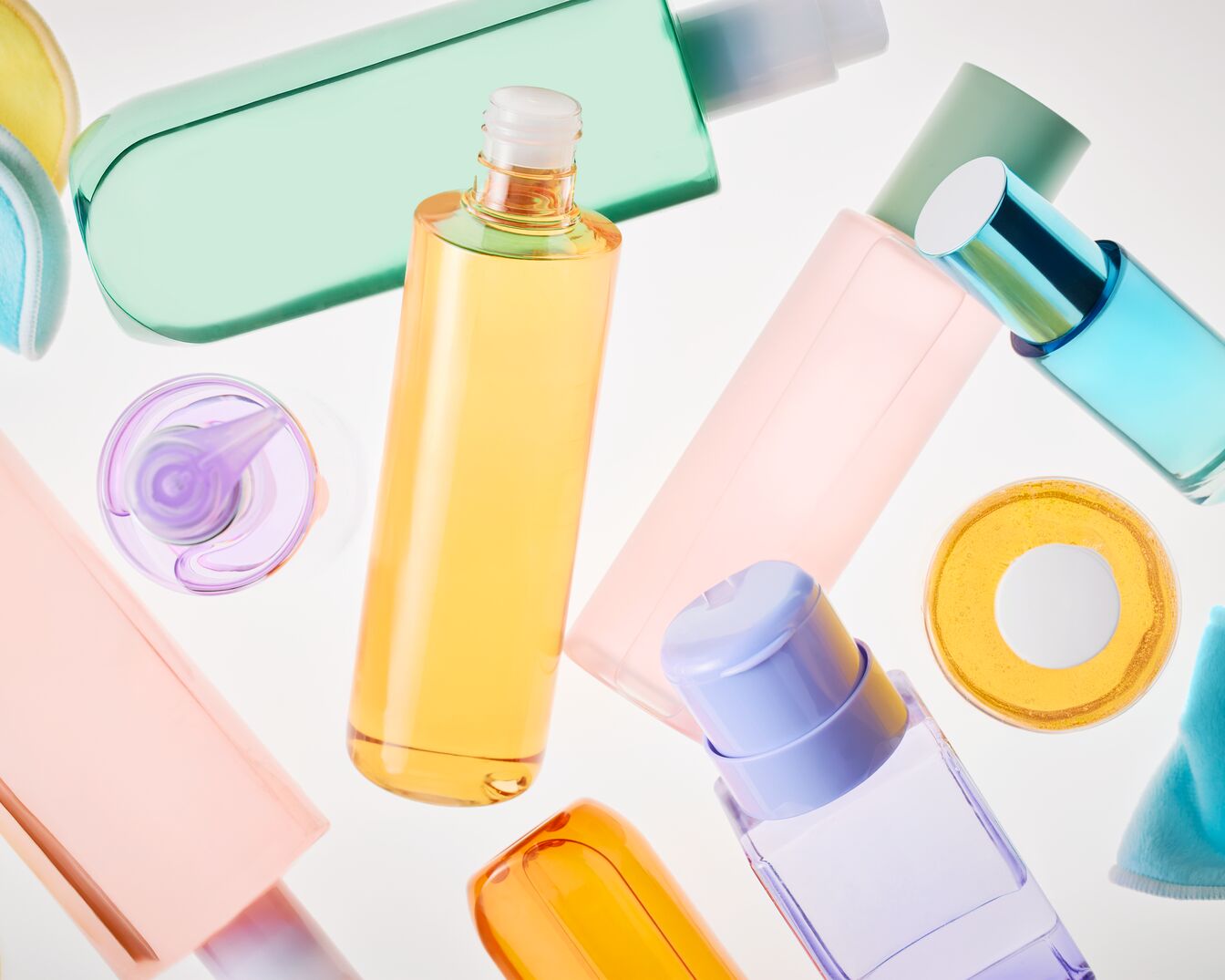Verschiedene Hautpflegeprodukte in Pastellfarben, darunter Flaschen mit gelben, rosa, grünen und blauen Flüssigkeiten, sowie ein runder Behälter mit einer gelben Flüssigkeit und ein blauer Waschlappen.