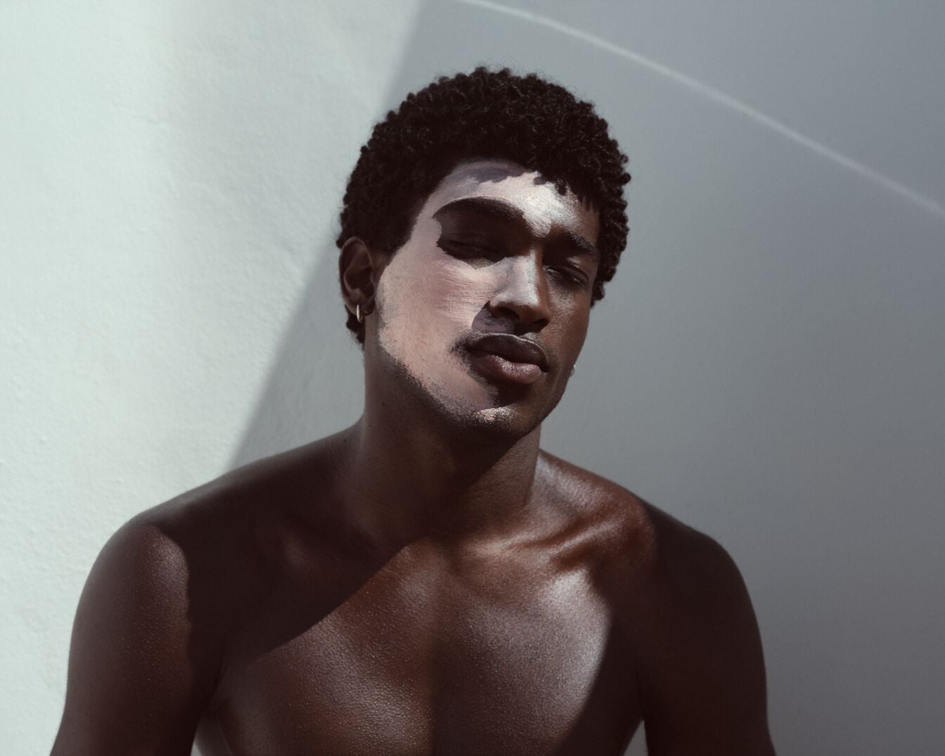 Ein Mann mit dunkler Haut und lockigem Haar hat eine weiße Gesichtsmaske aufgetragen und die Augen geschlossen.