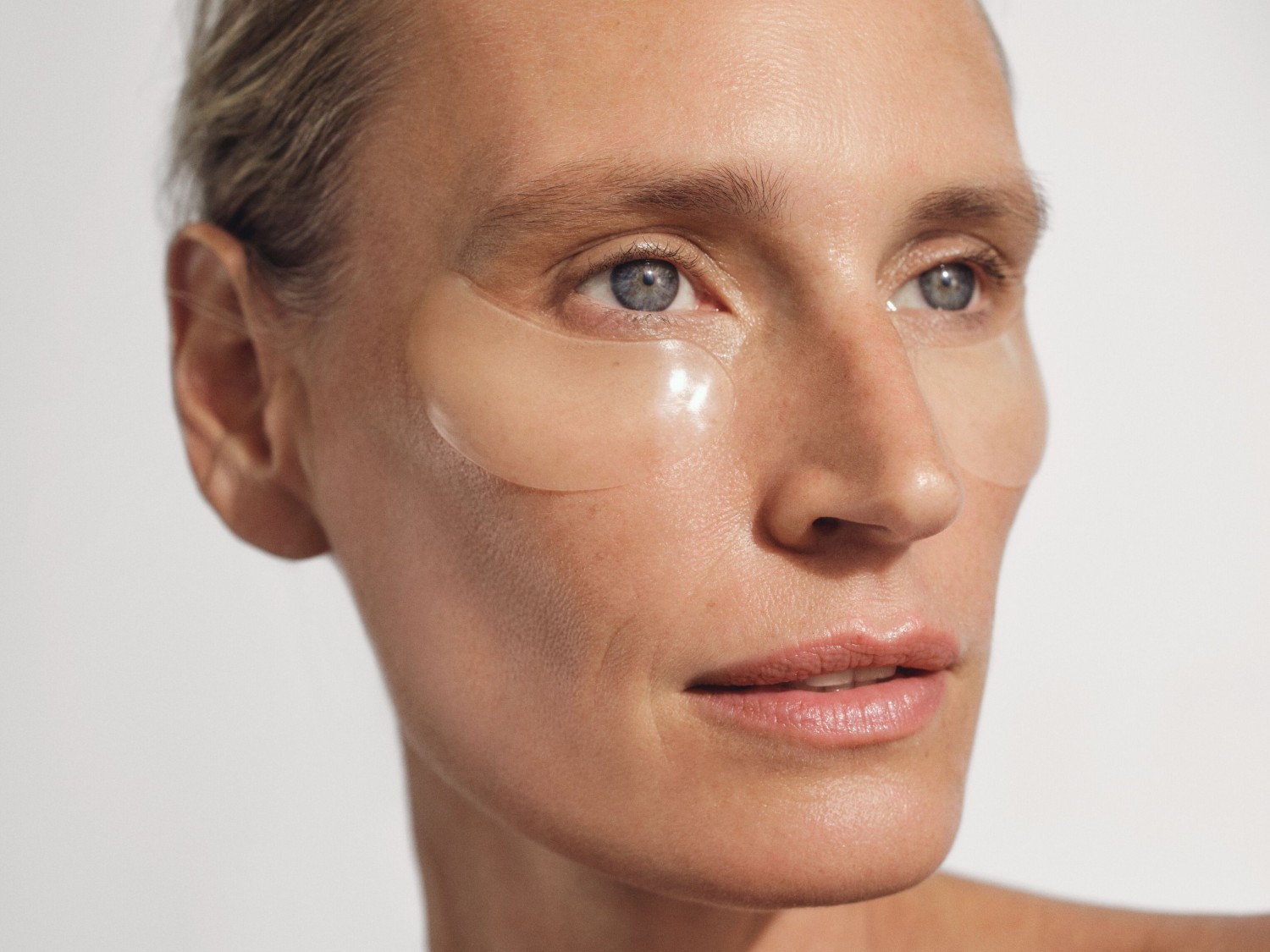 Nahaufnahme einer Frau mit hellblondem Haar und blauen Augen, die transparente Augenpads unter den Augen trägt, um die Haut zu pflegen.