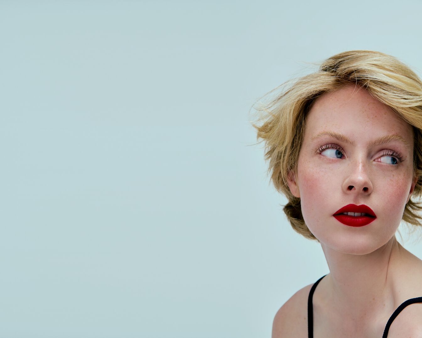 Porträt einer jungen Frau mit blonden, gewellten Haaren, roten Lippen und Sommersprossen, die über ihre Schulter in die Ferne blickt.