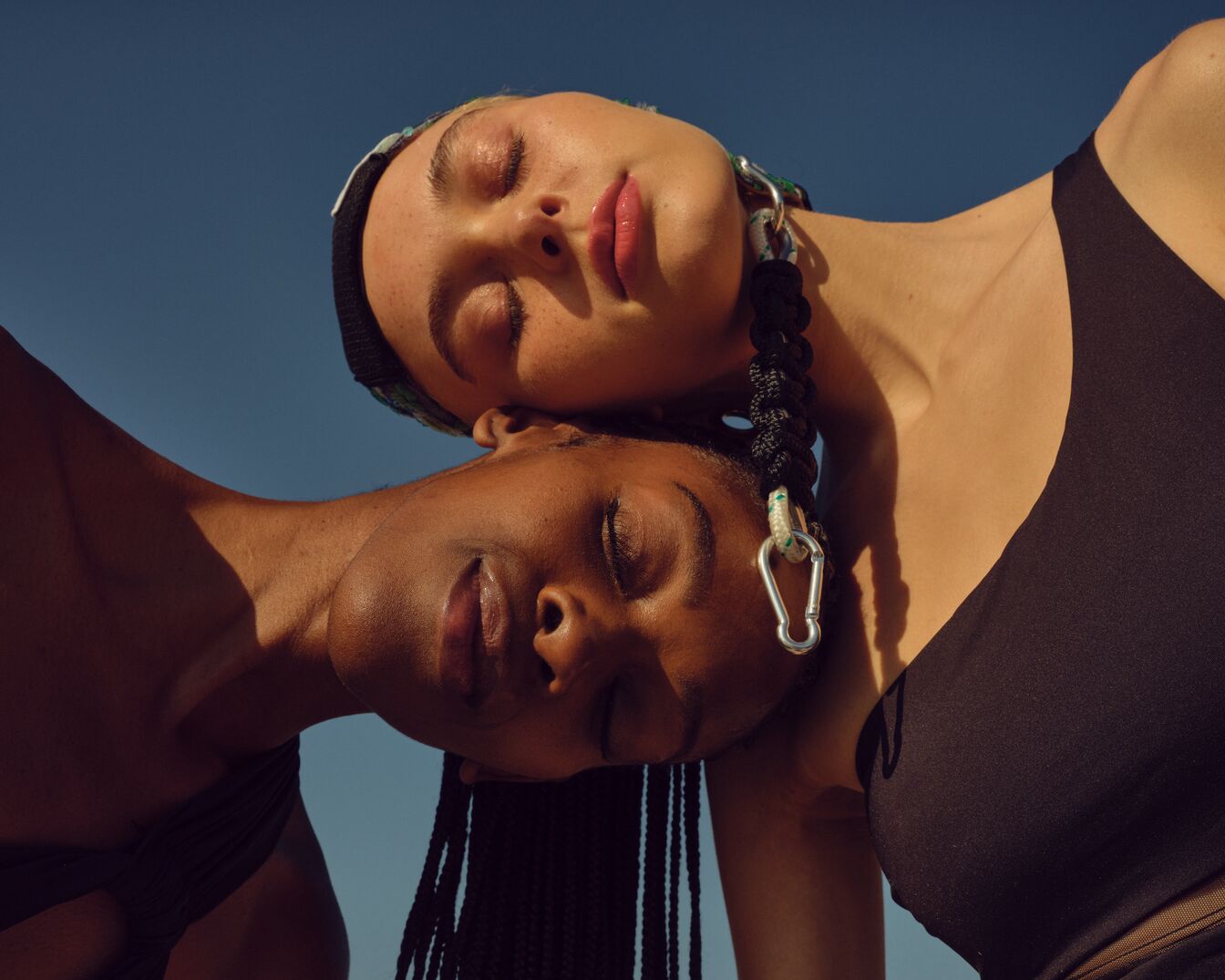 Zwei Frauen mit geschlossenen Augen, die sich gegenseitig die Köpfe anlehnen, wobei die obere eine gemusterte Kopfbedeckung und eine geflochtene Halskette mit silbernen Akzenten trägt, während die untere dunkle Zöpfe hat.