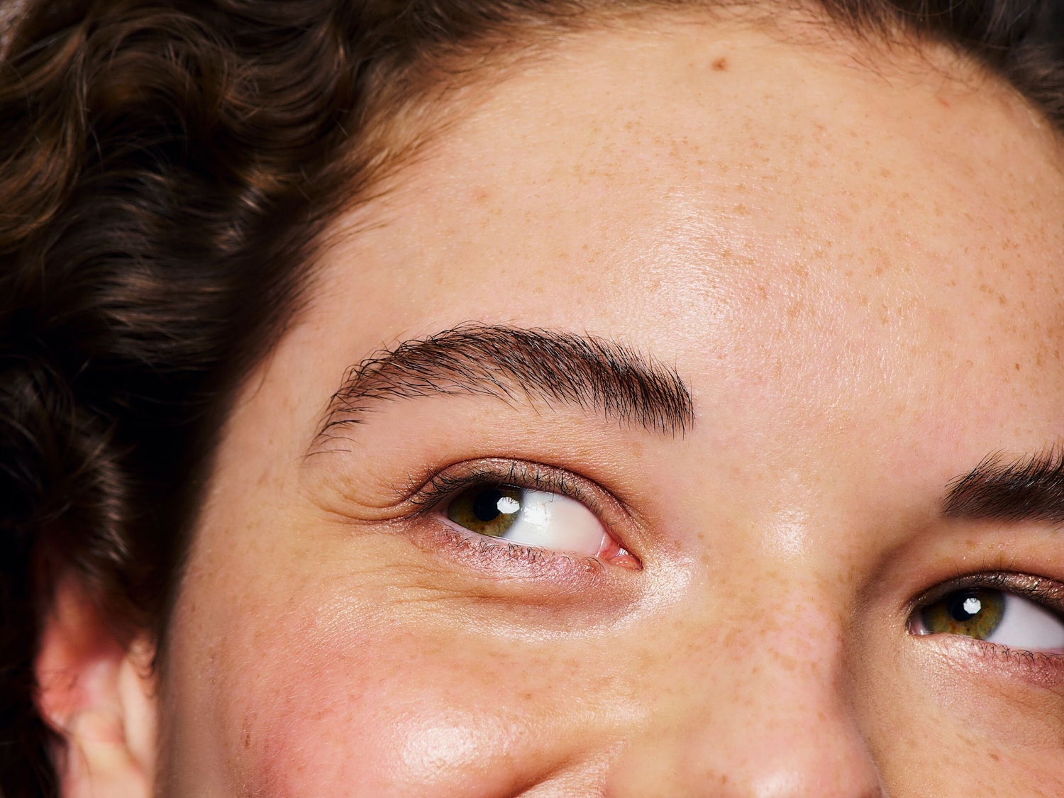 Nahaufnahme eines Gesichtes mit Sommersprossen, braunen Augen und dunklen, gewellten Haaren, die Stirnpartie betonend.