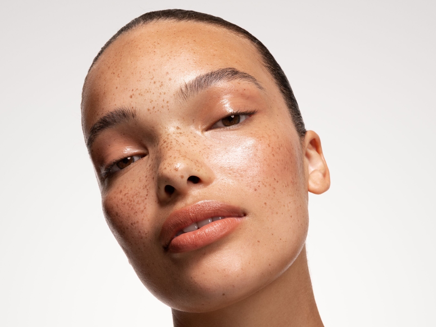 Nahaufnahme eines jungen Frauengesichts mit Sommersprossen, glänzender Haut und dezentem Make-up in Nude-Tönen.