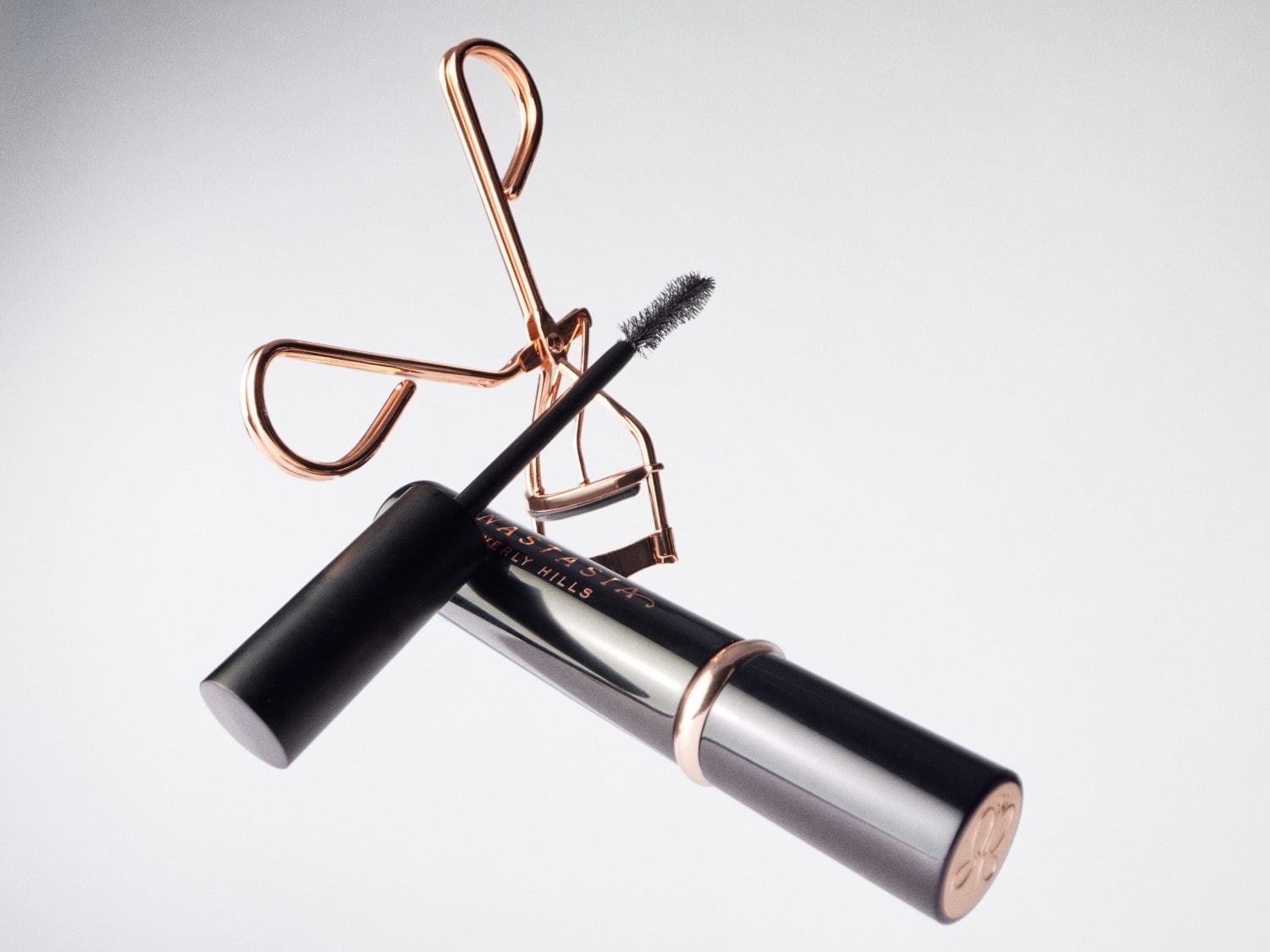 Eine schwarze Mascara von Anastasia Beverly Hills liegt neben einer goldenen Wimpernzange.