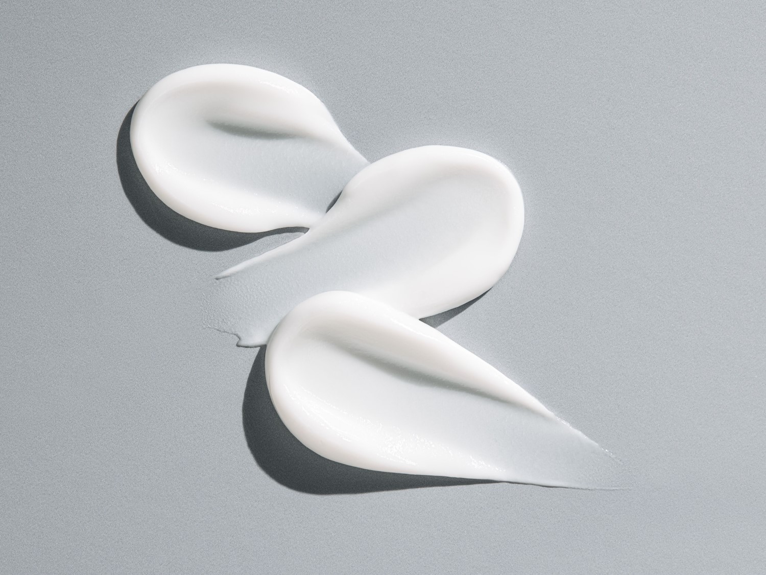 Tres trazos de crema blanca y brillante sobre un fondo gris claro, mostrando una textura suave y rica.