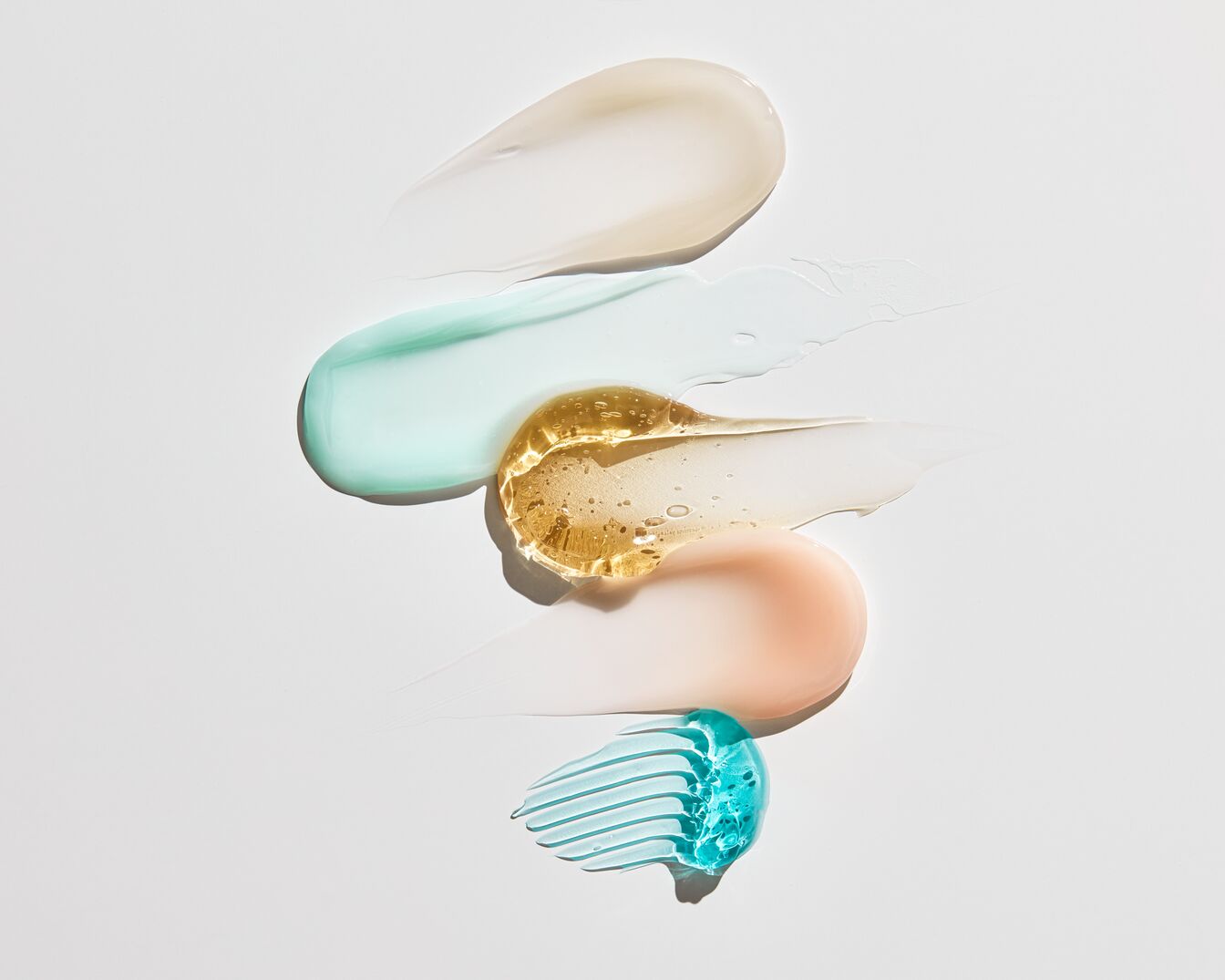 Na białym tle widoczne są próbki kosmetyków o różnych konsystencjach i kolorach, od kremowych po żelowe, w odcieniach beżu, zieleni, złota, różu i błękitu.