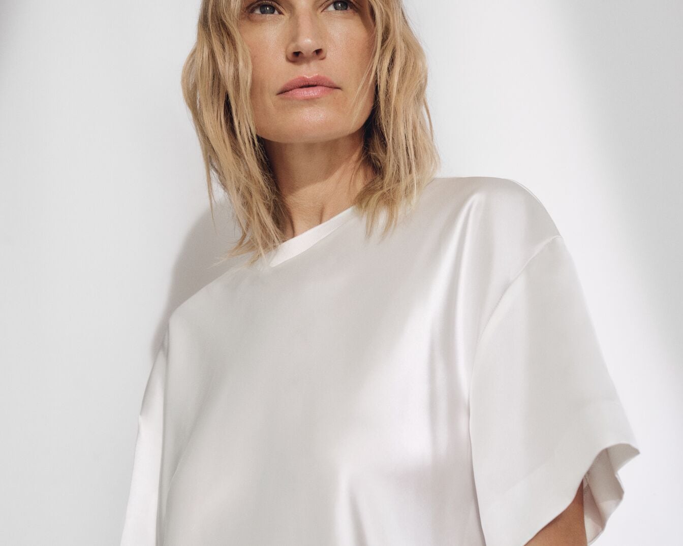 Portret kobiety z blond włosami, ubranej w białą, satynową bluzkę z krótkim rękawem, na jasnym tle.