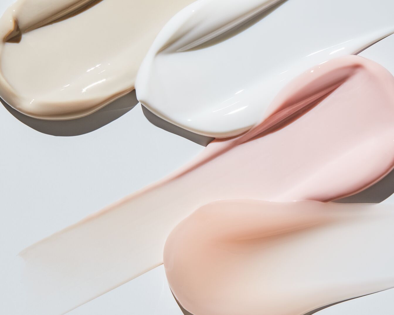 Kremy w odcieniach beżu, bieli i różu prezentują swoją gładką konsystencję na białym tle, idealne do pielęgnacji skóry.