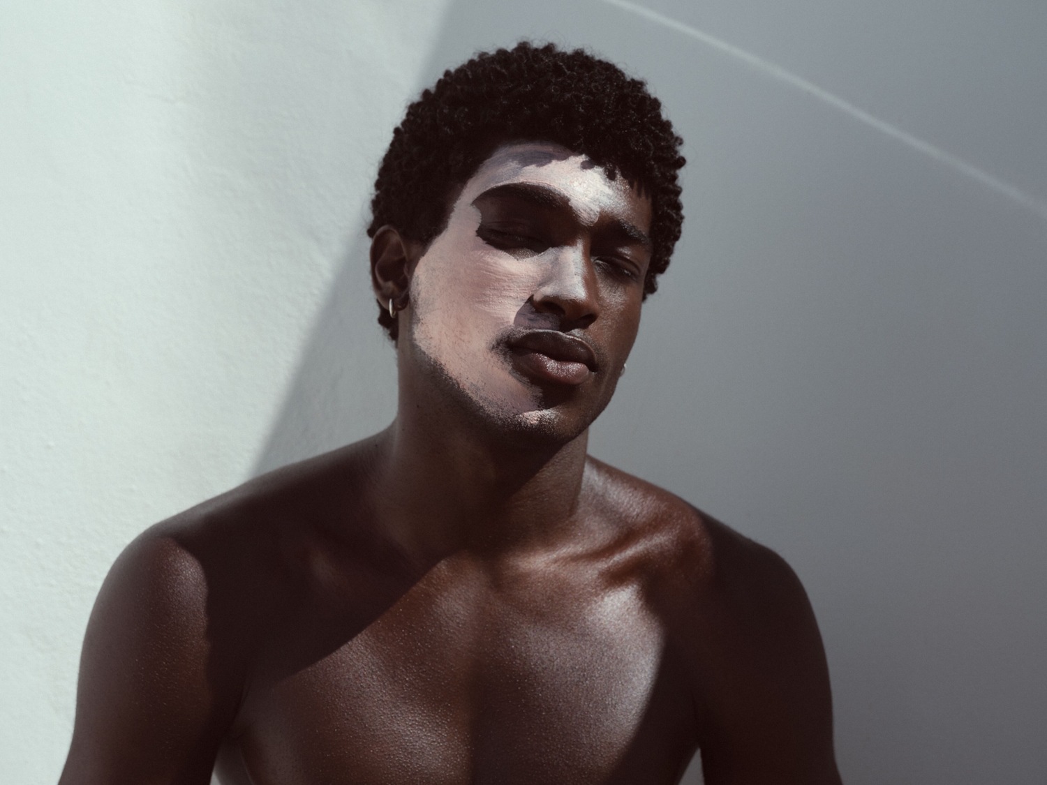 Mężczyzna o ciemnej karnacji z zamkniętymi oczami ma nałożoną maskę na część twarzy, stojąc na tle jasnej ściany.