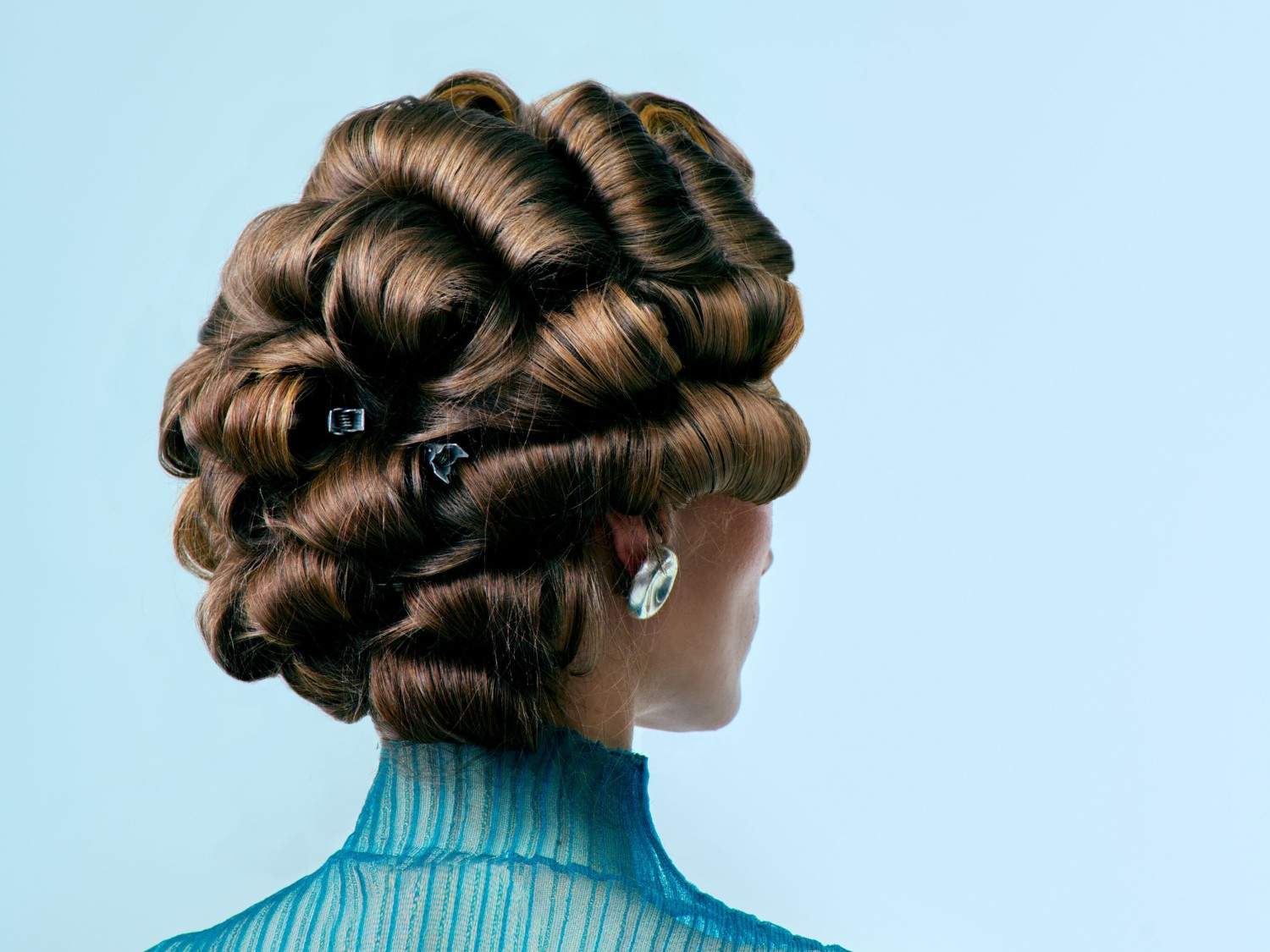 Bruin haar kunstig opgestoken in golvende rollen, vastgezet met speldjes, gecombineerd met een doorschijnende blauwe top en zilveren oorbellen tegen een lichtblauwe achtergrond.