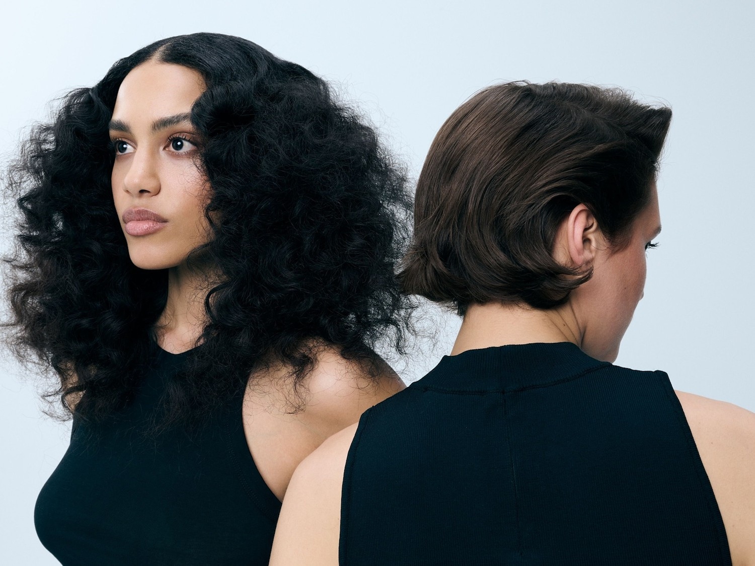 Twee vrouwen met verschillende haartypes, de een met lang, krullend zwart haar en de ander met kort, steil bruin haar, dragen zwarte mouwloze tops en staan rug aan rug.