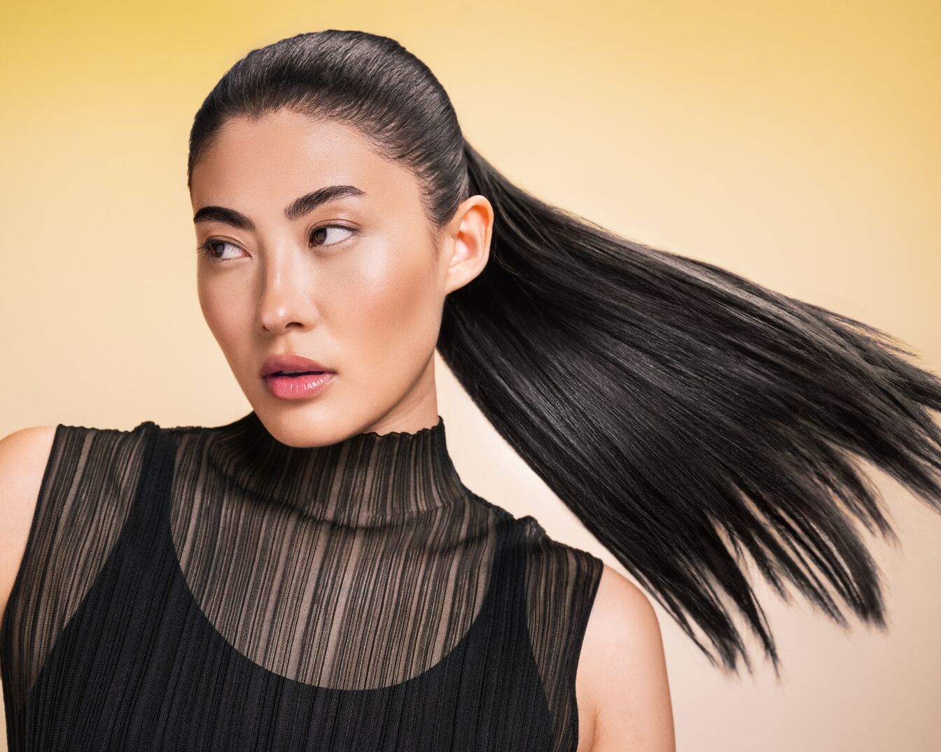 Een vrouw met glanzend, donker haar in een strakke paardenstaart, gekleed in een zwarte top, kijkt weg tegen een zachtgele achtergrond.