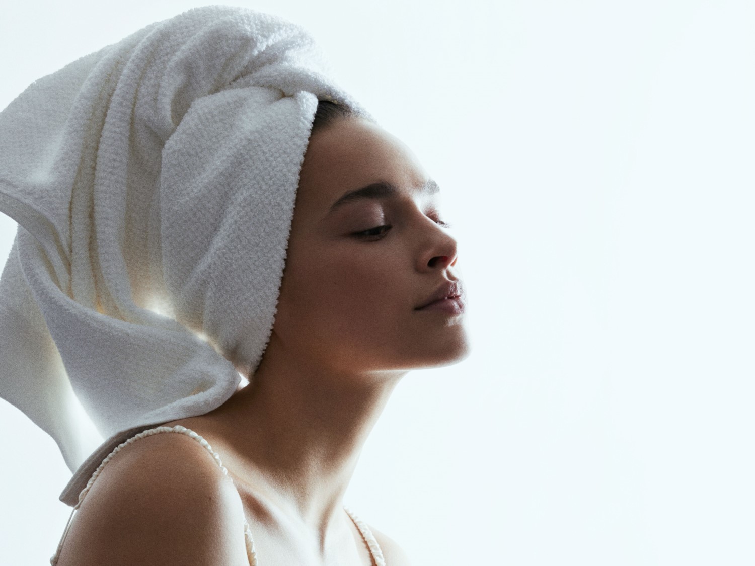 Profil kobiety z białym ręcznikiem na głowie, z zamkniętymi oczami, na jasnym tle, emanująca spokojem i świeżością.