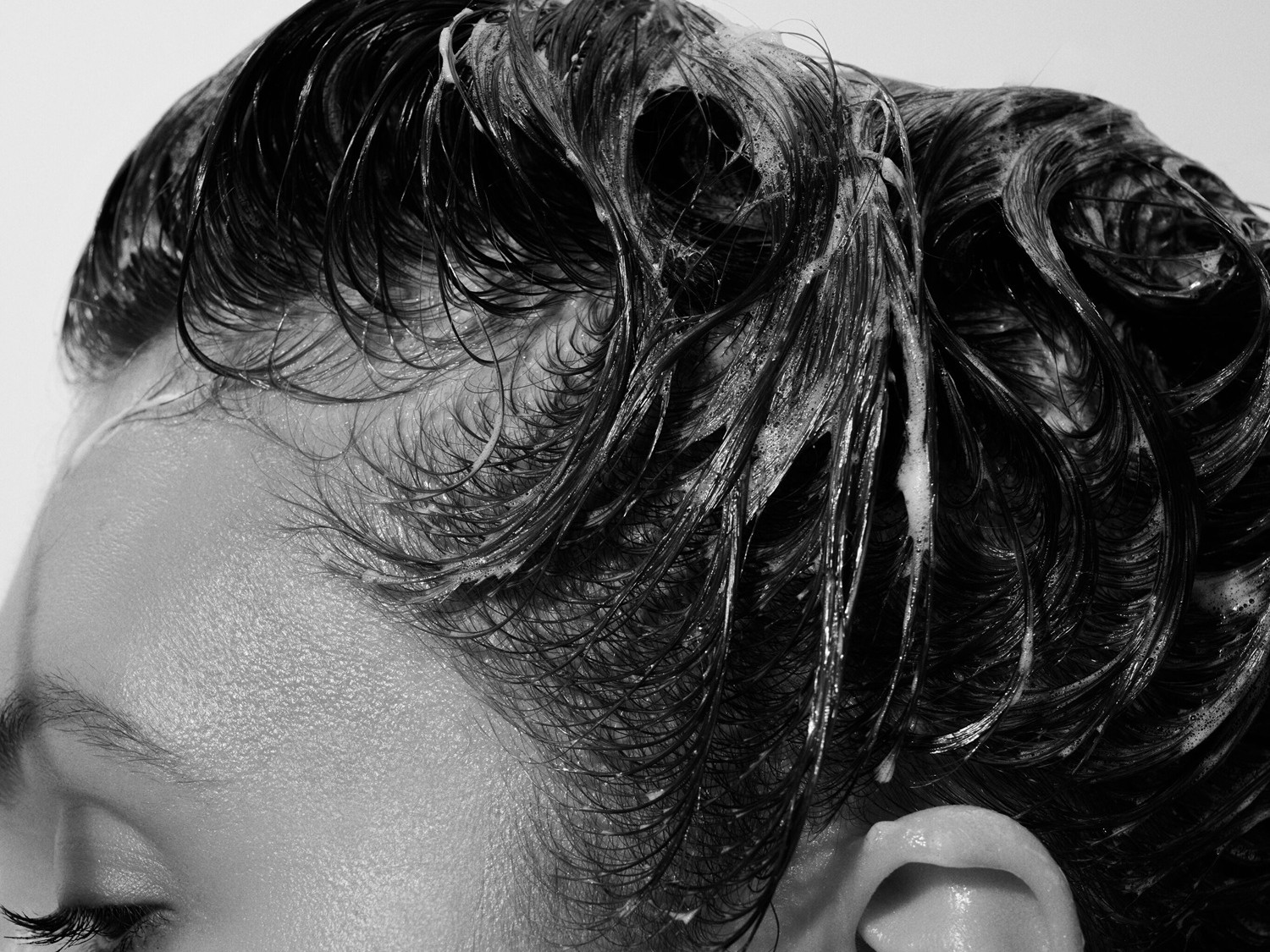 Una persona con i capelli scuri bagnati e insaponati, con schiuma bianca visibile tra le ciocche, che mostra l'efficacia del prodotto per la cura dei capelli.