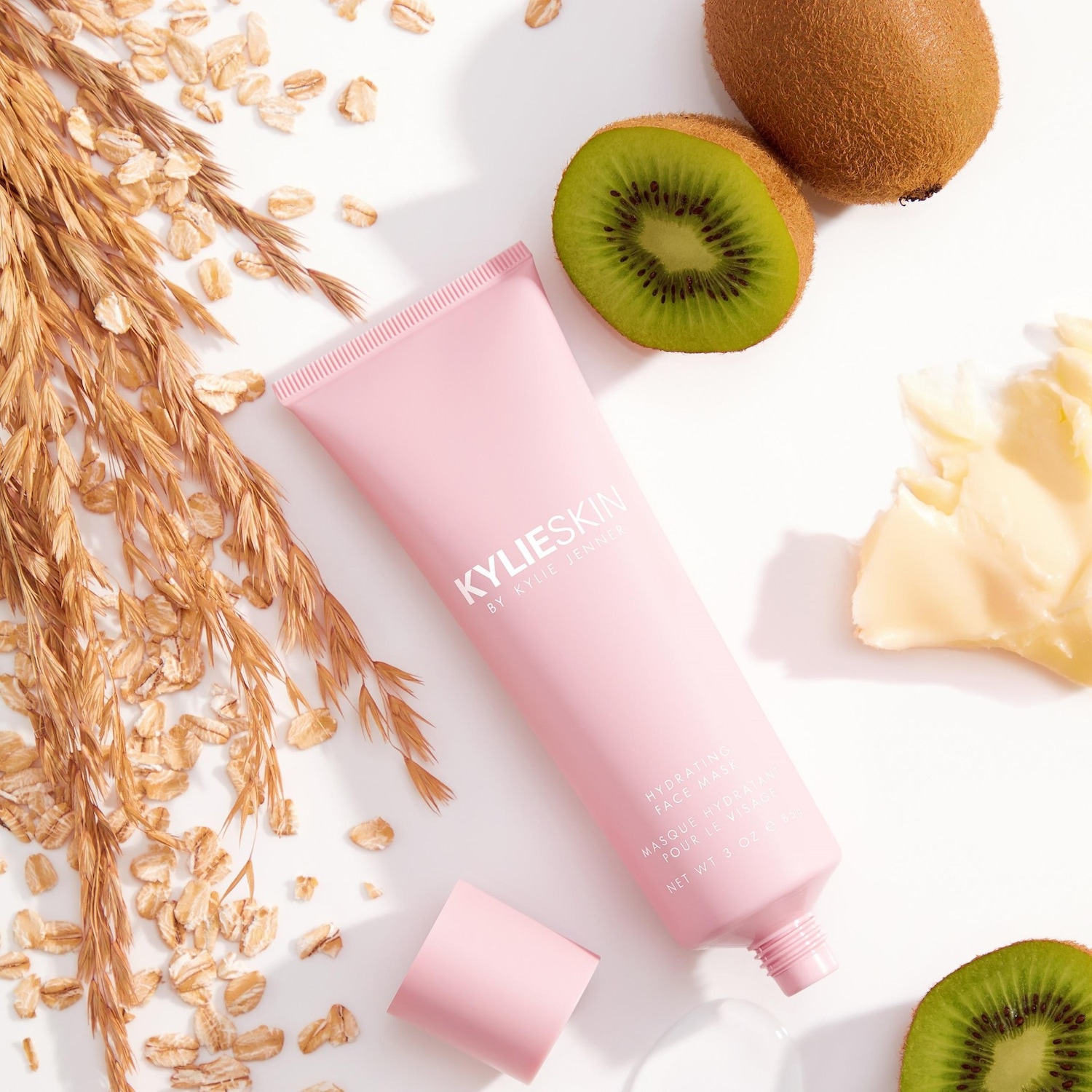 Le masque hydratant pour le visage Kylie Skin est présenté dans un tube rose pâle, entouré de kiwis frais, d'avoine et d'une touche de beurre pour une peau éclatante.