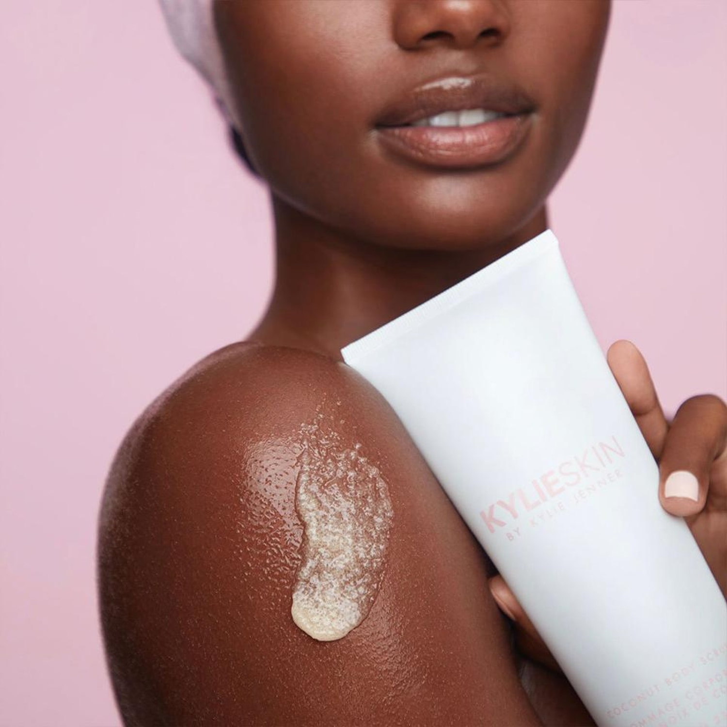Une femme à la peau foncée tient un tube blanc de gommage corporel Kylie Skin, avec une noisette de produit exfoliant sur son épaule, le tout sur un fond rose uni.