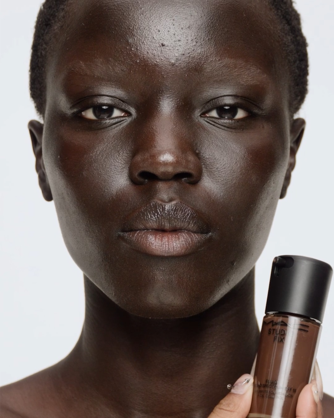 Een vrouw met een donkere huidskleur houdt een flesje MAC Studio Fix foundation vast, perfect passend bij haar teint, voor een egale en stralende look.