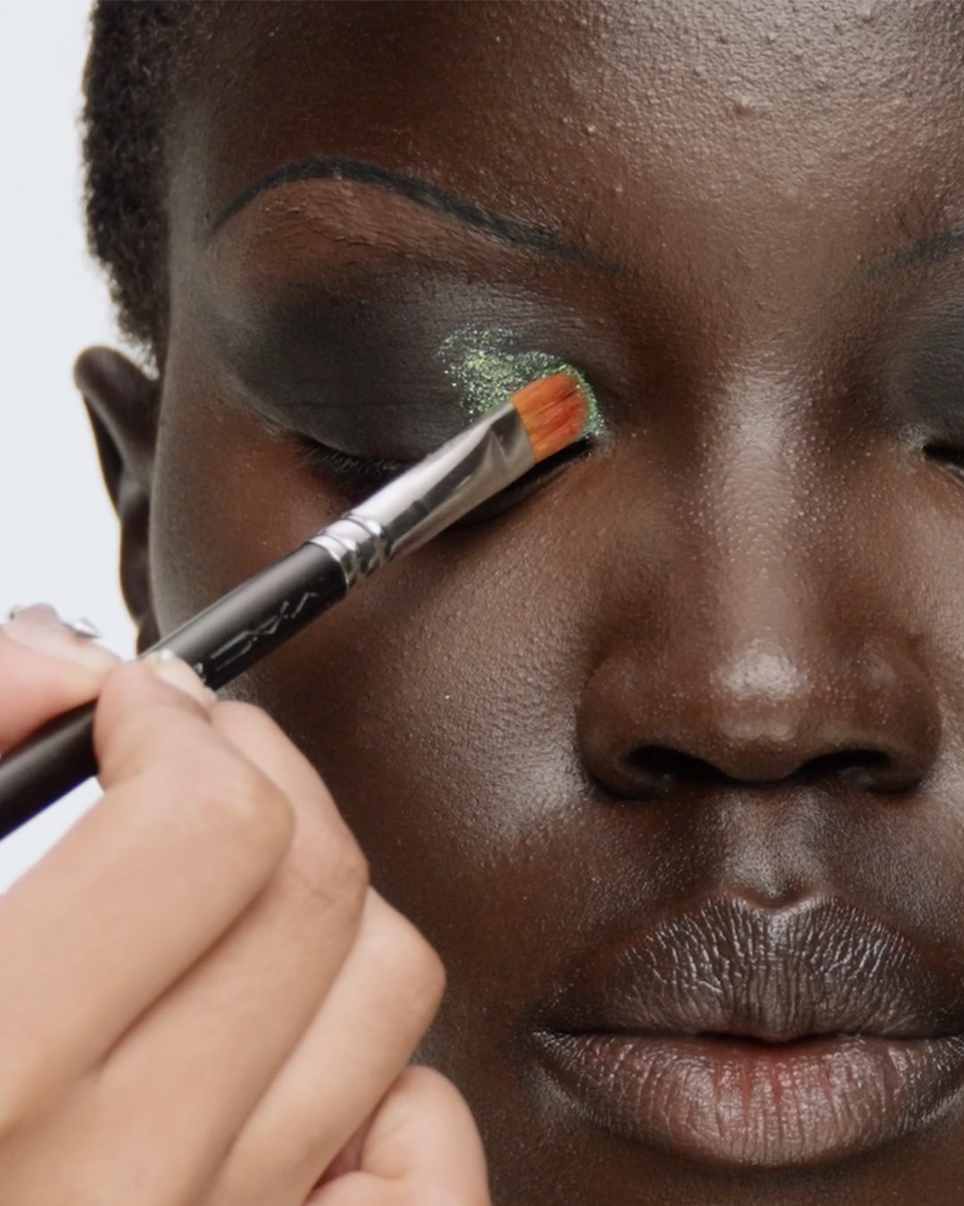 Een close-up van een donkerhuidige vrouw die groene glitteroogschaduw op haar ooglid aanbrengt met een make-upborstel.