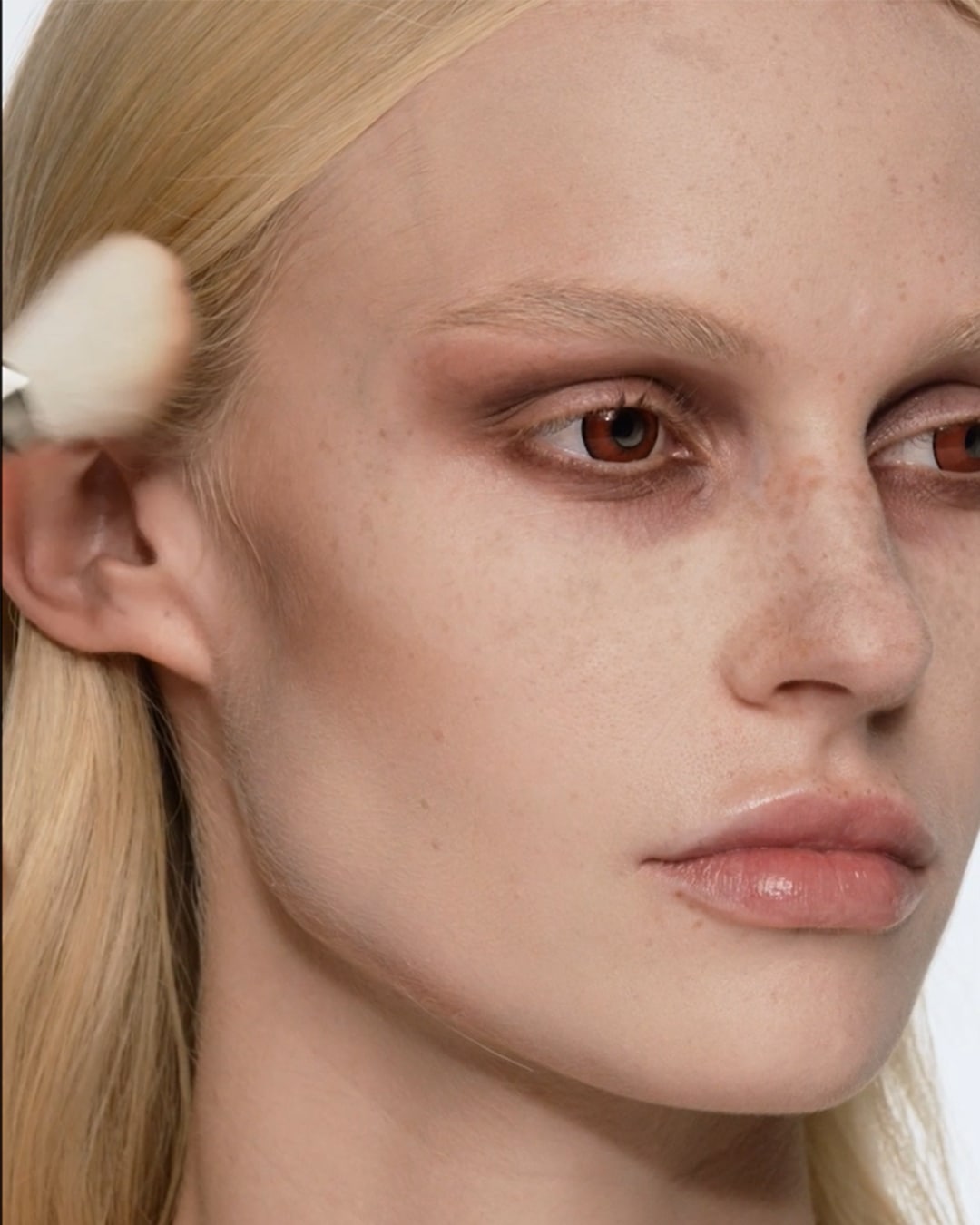 Close-up van een vrouw met blond haar en bruine oogschaduw die met een make-upborstel foundation aanbrengt op haar gezicht.