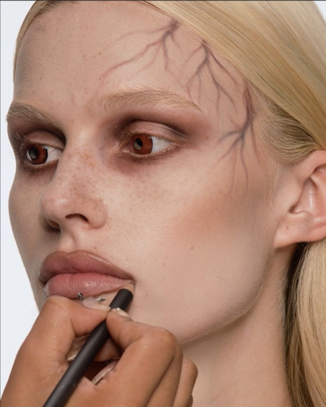 Een visagist brengt met een zwart potlood make-up aan op de lippen van een model met een griezelige Halloween-look, inclusief aderen op het gezicht.
