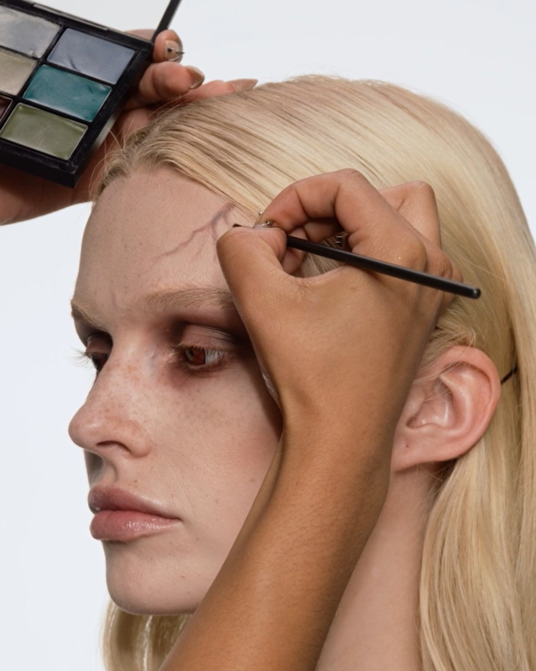 Een visagist gebruikt een penseel en een palet met groene en grijze tinten om een litteken op het voorhoofd van een blond model te tekenen.