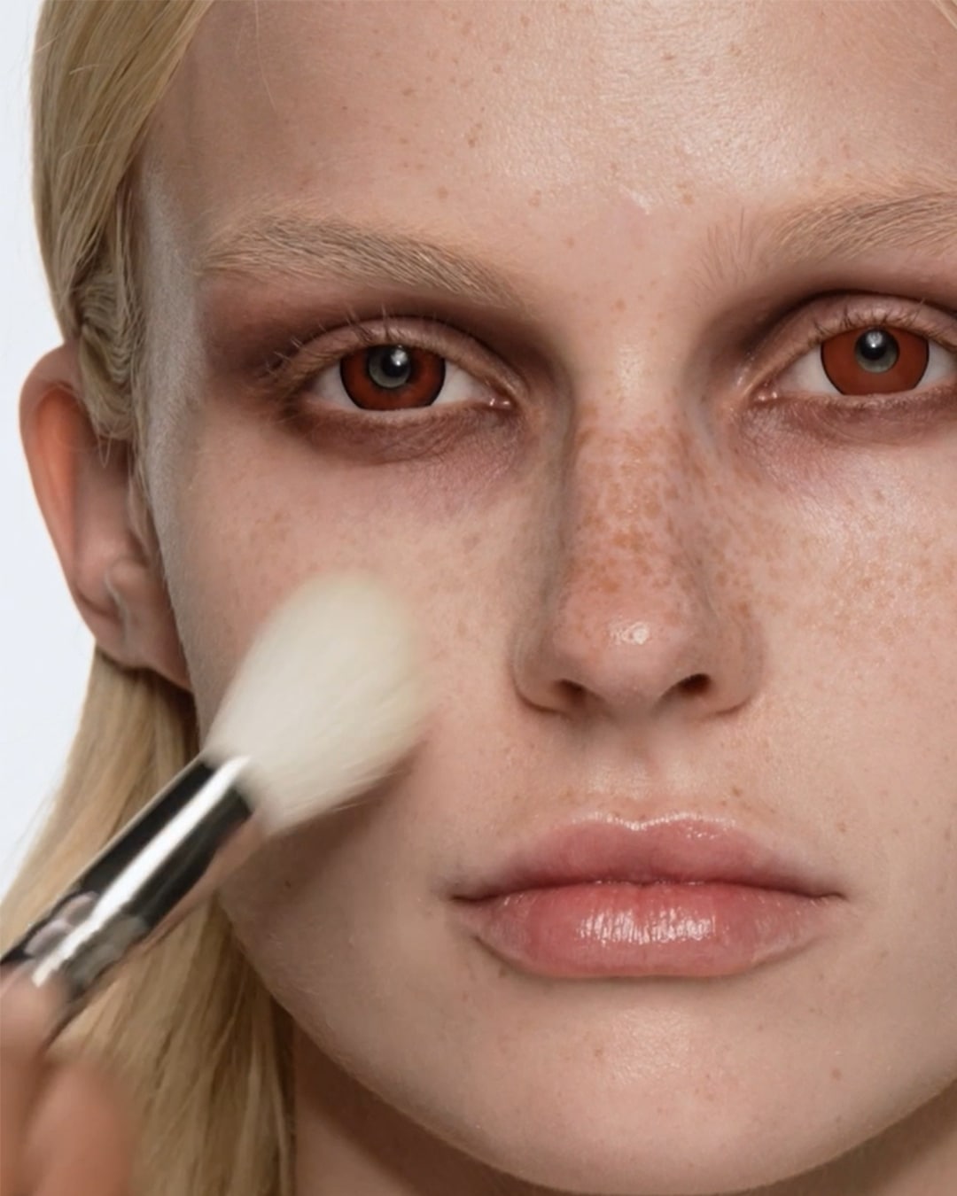 Een vrouw met een lichte huid en sproeten brengt make-up aan met een witte borstel, wat zorgt voor een zachte en egale teint.