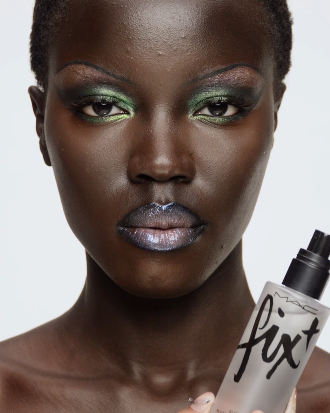 Een vrouw met een donkere huidskleur heeft groene oogschaduw en glanzende lippen, terwijl ze een fles MAC Fix+ vasthoudt.