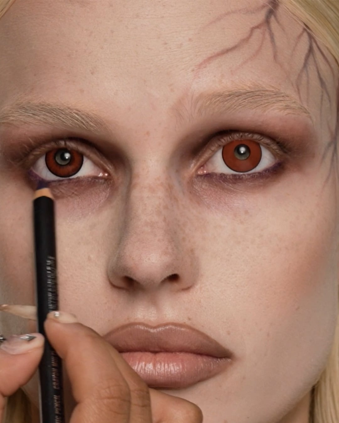 Een close-up van een vrouw met bruine ogen en een bleke huid, terwijl een hand een zwart oogpotlood aanbrengt om een griezelige Halloween-look te creëren.