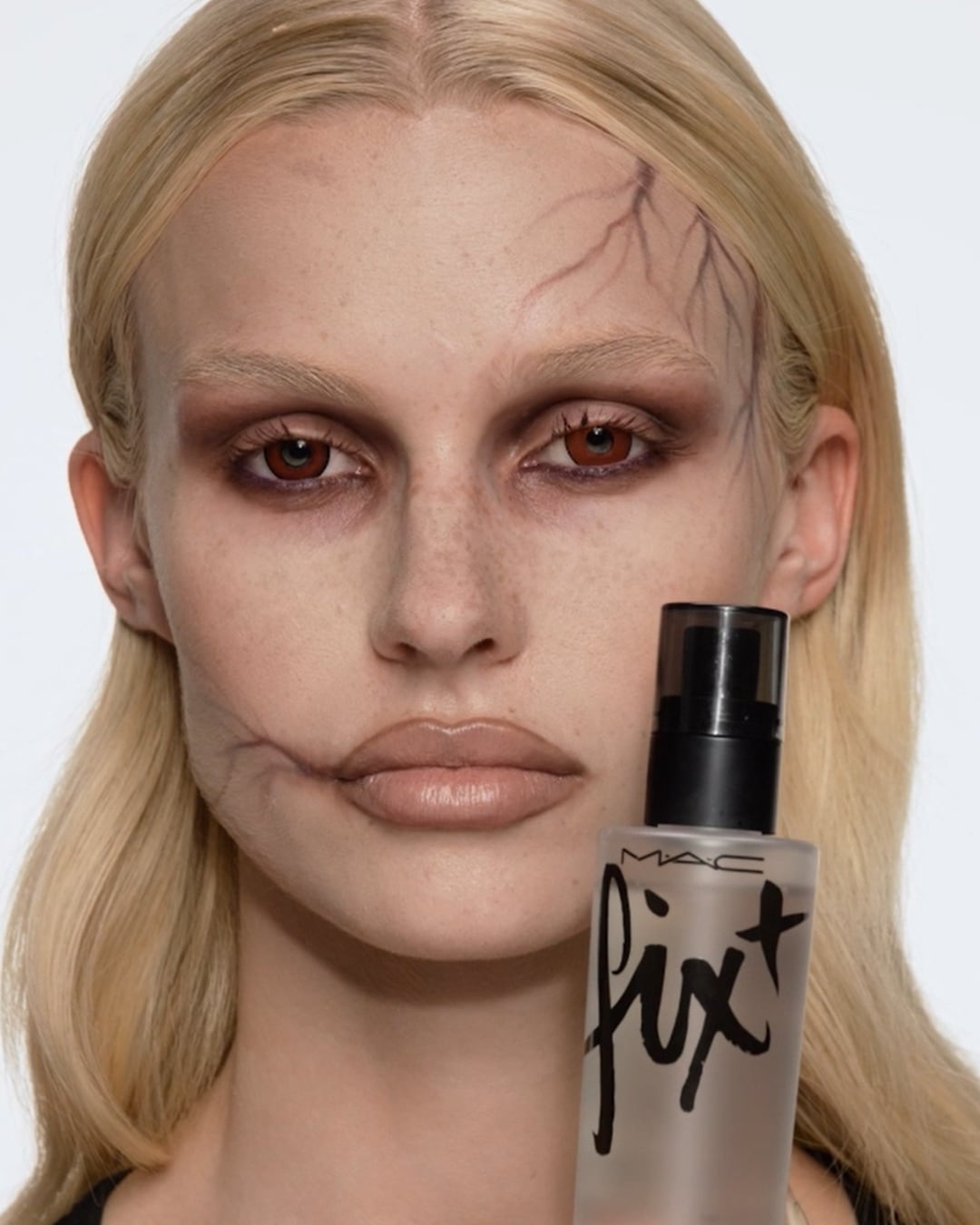 Een vrouw met Halloween-make-up houdt een MAC Fix+ setting spray vast, perfect om je griezelige look de hele nacht te fixeren.