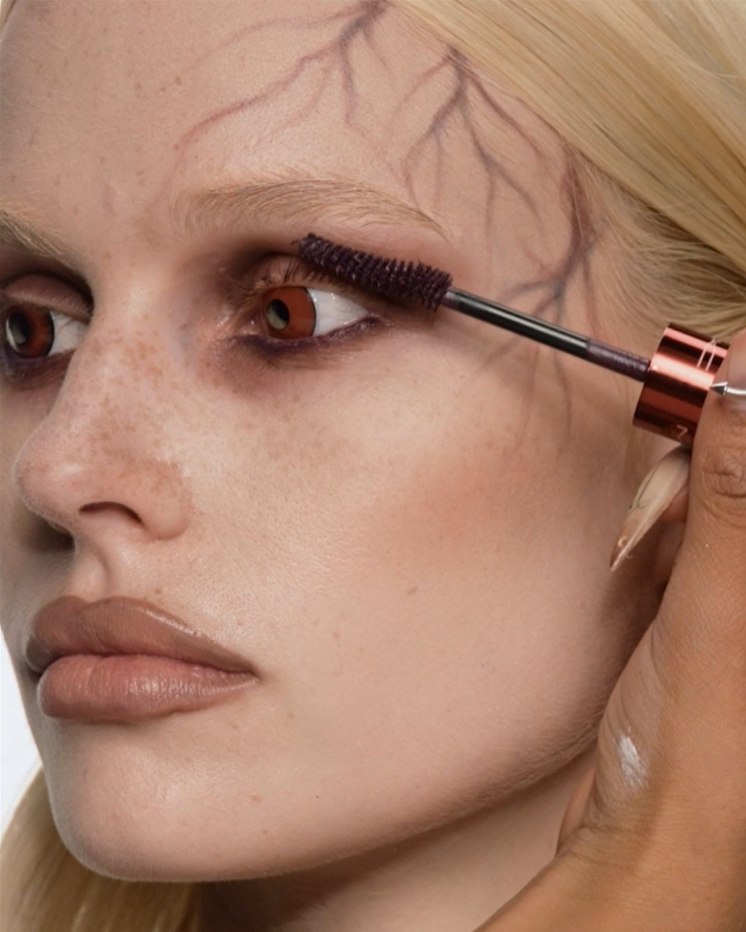 Een close-up van een vrouw met blond haar die mascara aanbrengt, met een artistiek ontwerp van takken op haar voorhoofd.