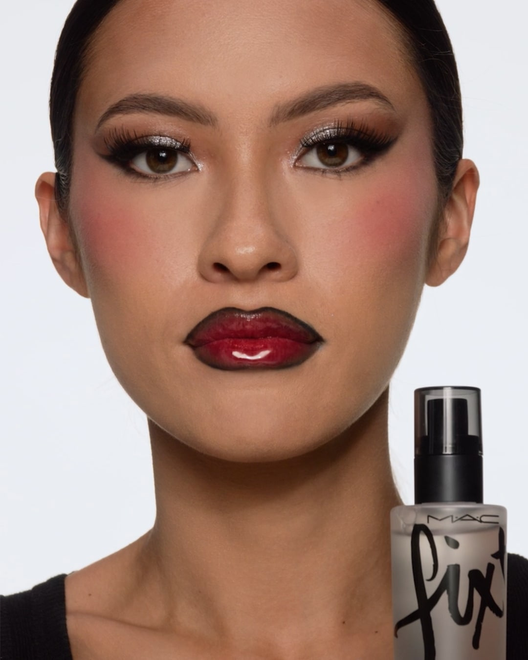 Een close-up van een vrouw met een gedurfde make-up look, inclusief zilveren oogschaduw, rode blush en een ombre lip met rode en zwarte tinten, met een MAC Fix+ setting spray in de voorgrond.
