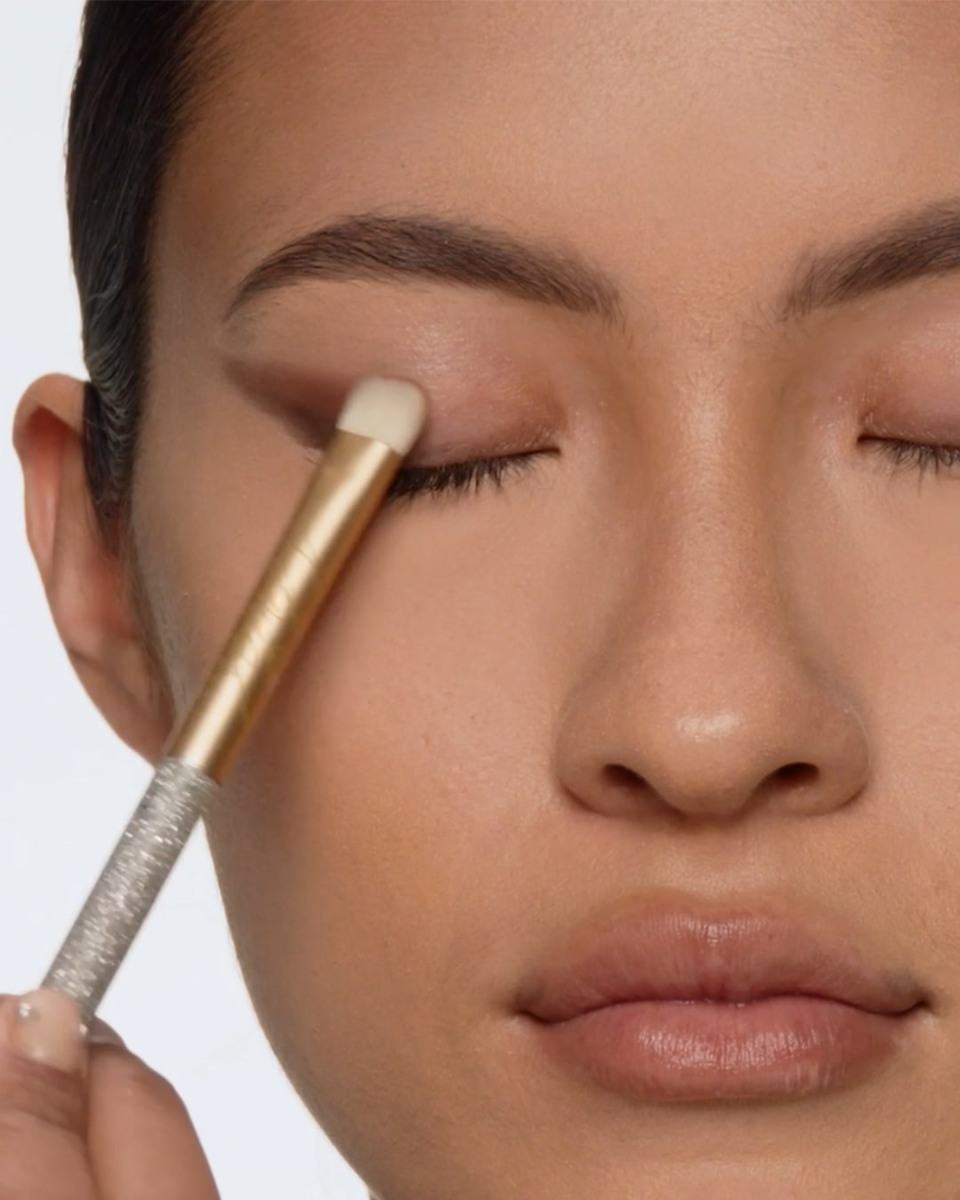 Een vrouw brengt oogschaduw aan met een gouden kwast, haar ogen gesloten voor een nauwkeurige applicatie.