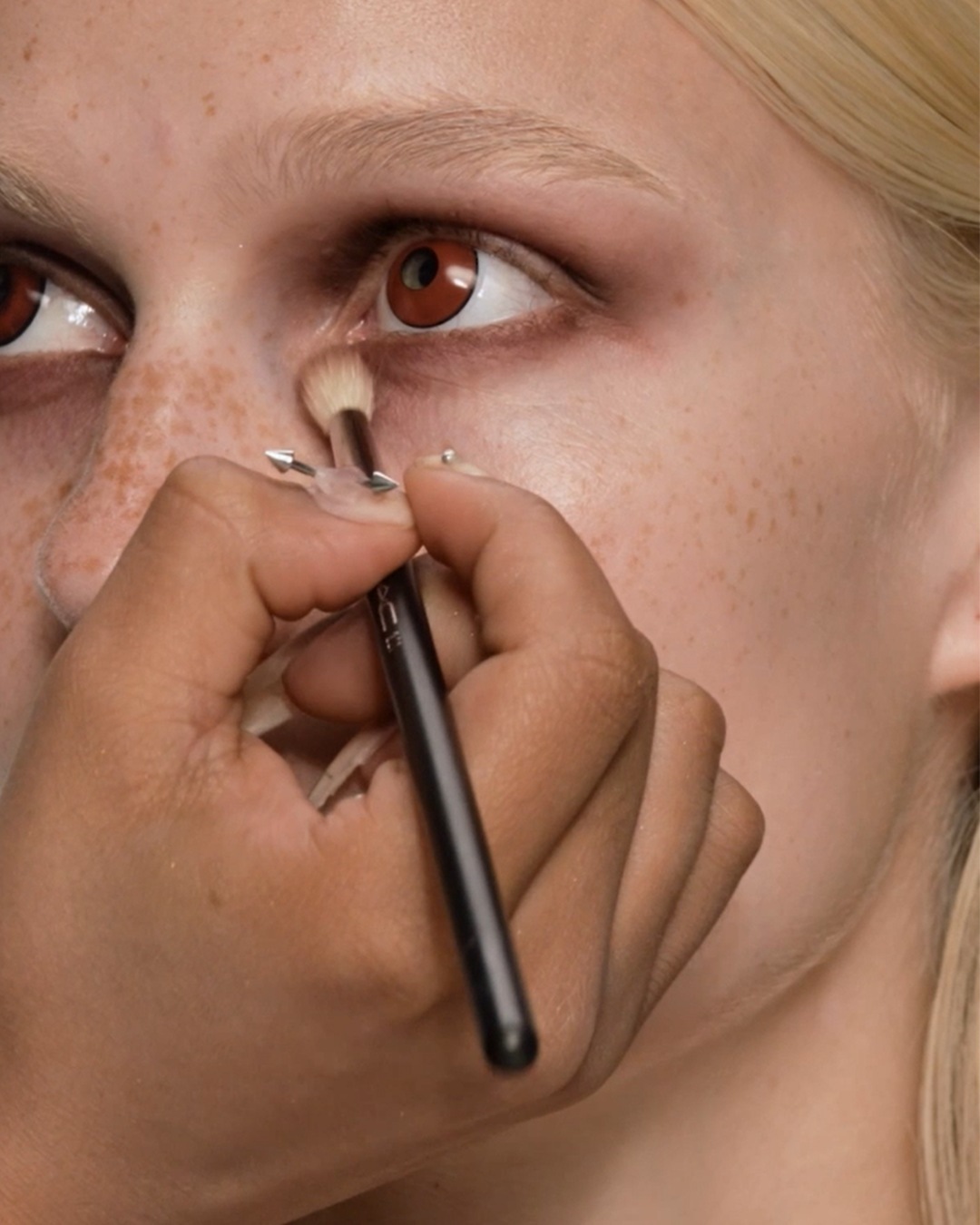 Een visagist brengt met een kleine make-upborstel bruine oogschaduw aan onder het oog van een model met een lichte huid en bruine ogen.