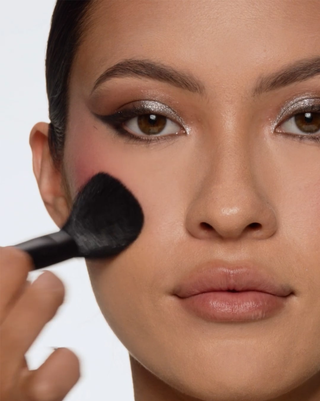 Een vrouw brengt met een zwarte make-upborstel roze blush aan op haar wang, haar ogen zijn opgemaakt met zilveren oogschaduw en zwarte eyeliner.