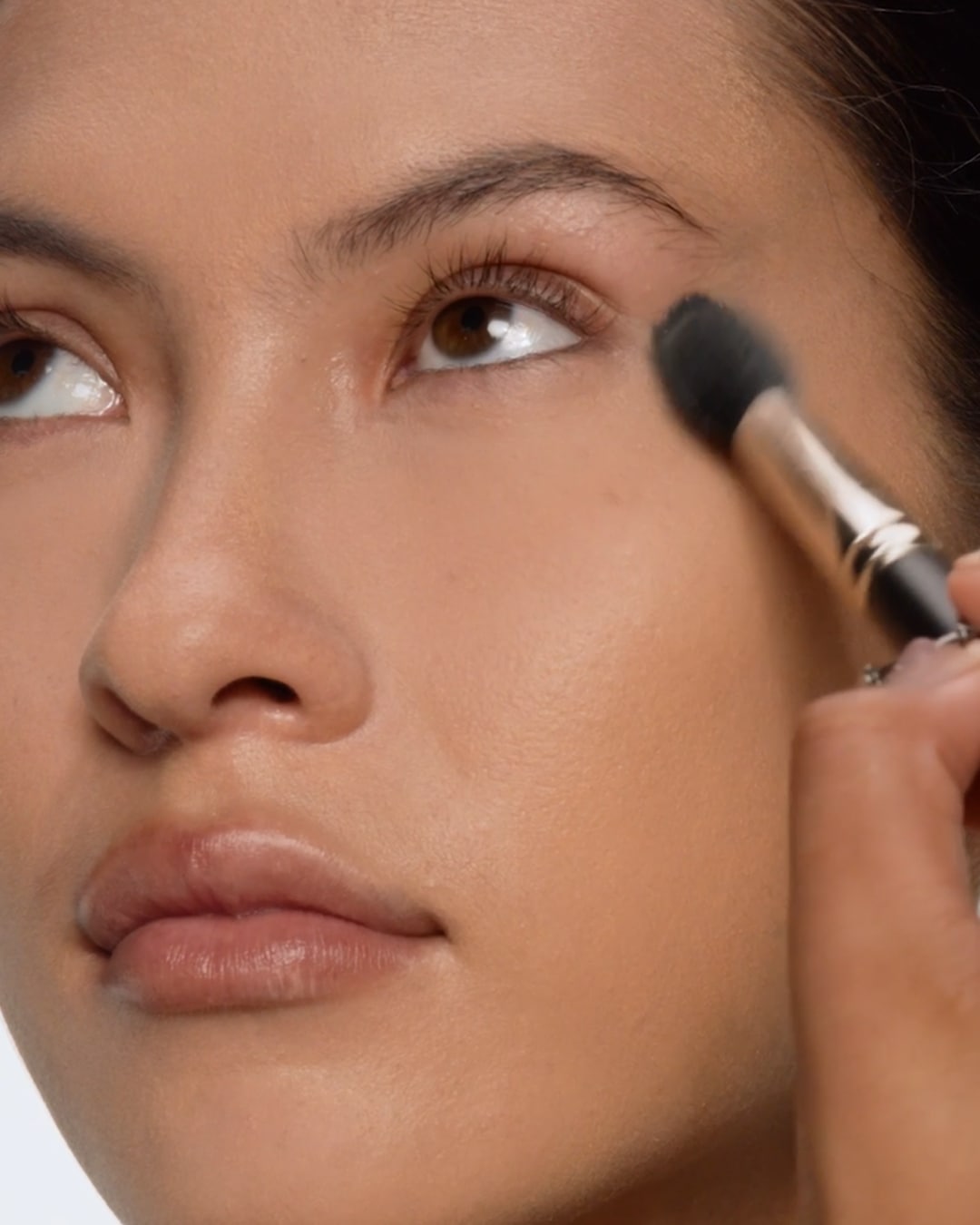 Een vrouw brengt make-up aan met een zwarte borstel onder haar oog, waarbij de borstel een gouden handvat heeft.