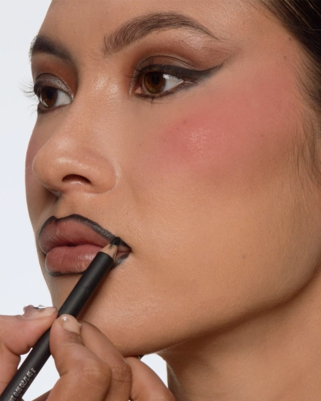 Een close-up van een vrouw die haar lippen omlijnt met een zwart lippotlood, haar make-up is compleet met een winged eyeliner en roze blos.