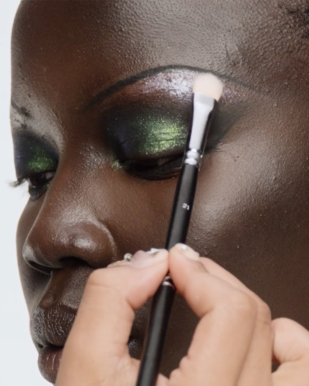 Een close-up van een persoon met een donkere huidskleur die oogschaduw aanbrengt met een make-upborstel, waarbij de groene en zilveren tinten van de oogschaduw worden benadrukt.