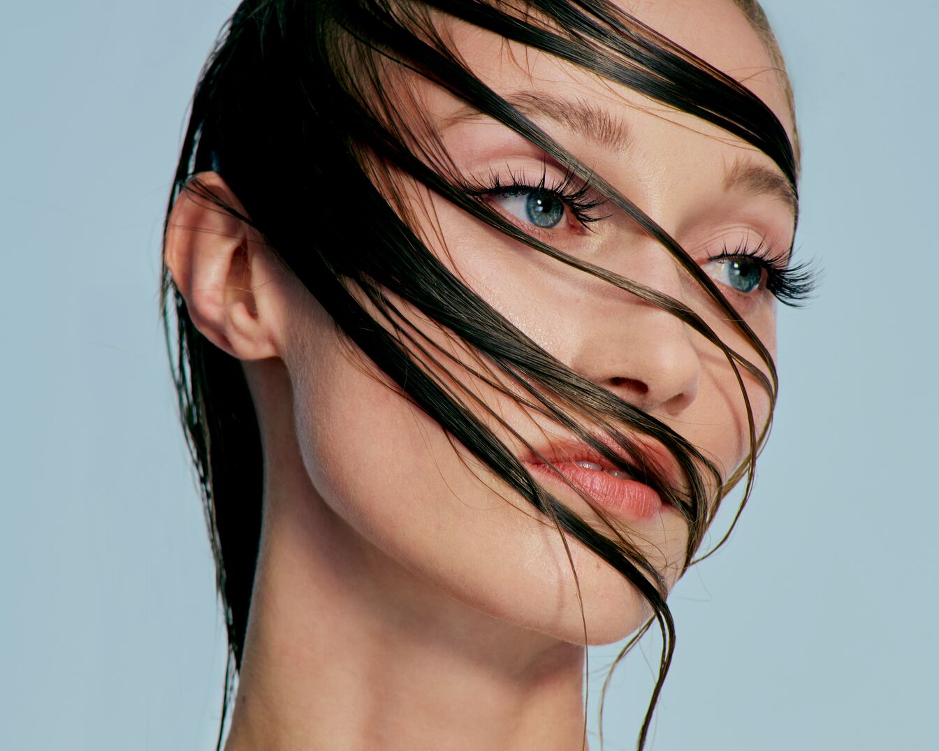 Close-up van een vrouw met nat, donker haar dat over haar gezicht valt, blauwe ogen en zachte lippenstift tegen een lichtblauwe achtergrond.
