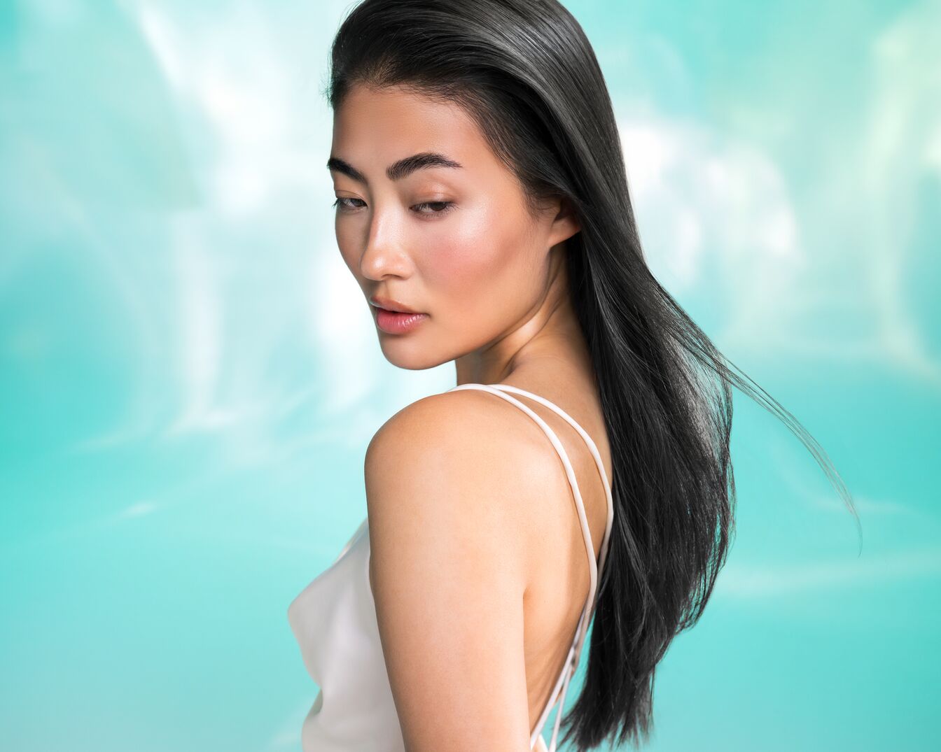 Een vrouw met lang, donker haar en een witte top kijkt over haar schouder tegen een zachtblauwe achtergrond.