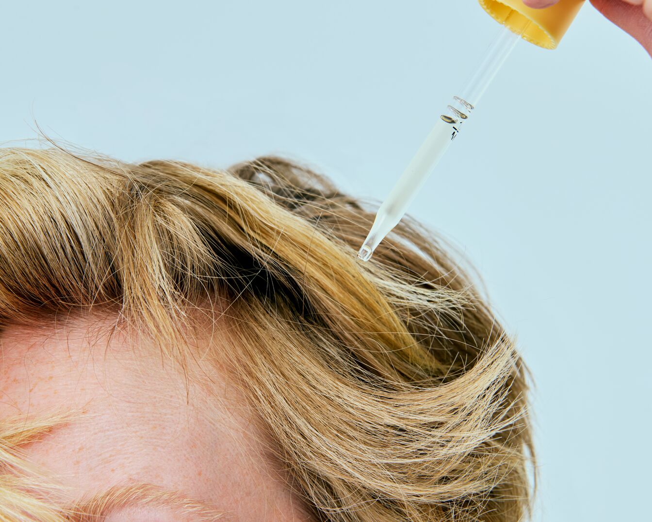 Een heldere vloeistof wordt met een glazen pipet op blond haar aangebracht, wat de essentie van haarverzorging en verjonging vastlegt.