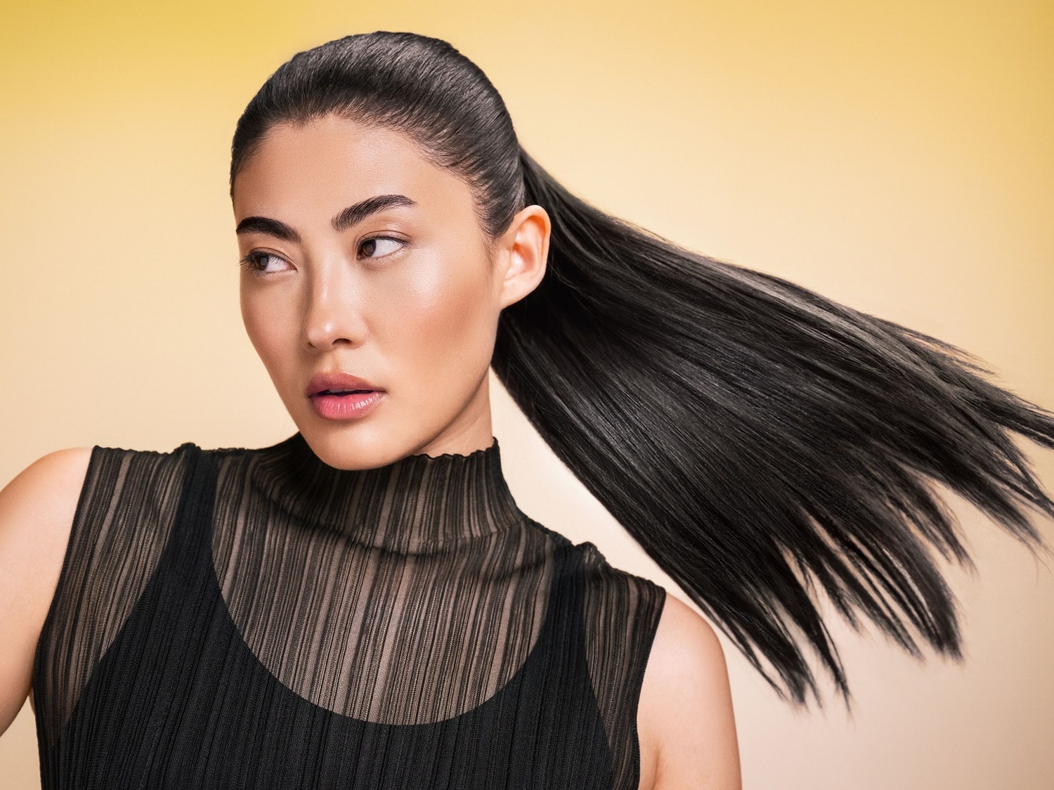 Primo piano di una donna asiatica con lunghi capelli neri raccolti in una coda di cavallo, che indossa un top nero a collo alto e guarda di lato su uno sfondo giallo.