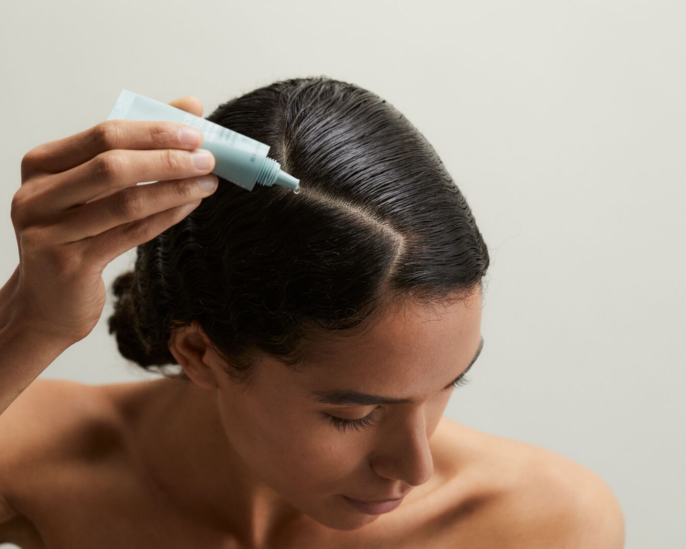 Una donna applica un trattamento sui capelli umidi con una confezione azzurra, concentrandosi sulla riga per nutrire il cuoio capelluto.