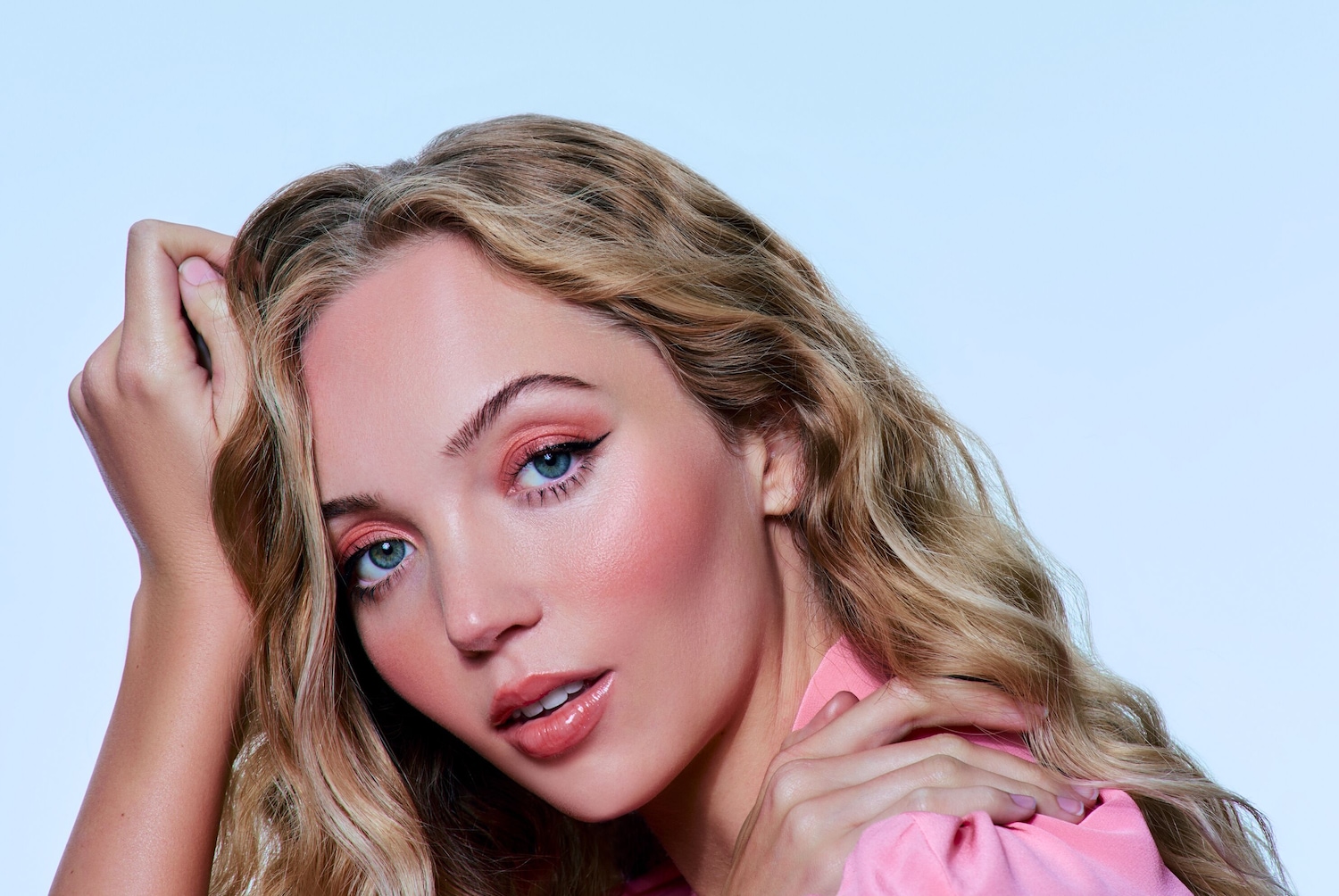 Nahaufnahme einer Frau mit gewelltem, blondem Haar, blauen Augen, zartem rosa Lidschatten und passendem Lippenstift, die einen rosa Blazer trägt.