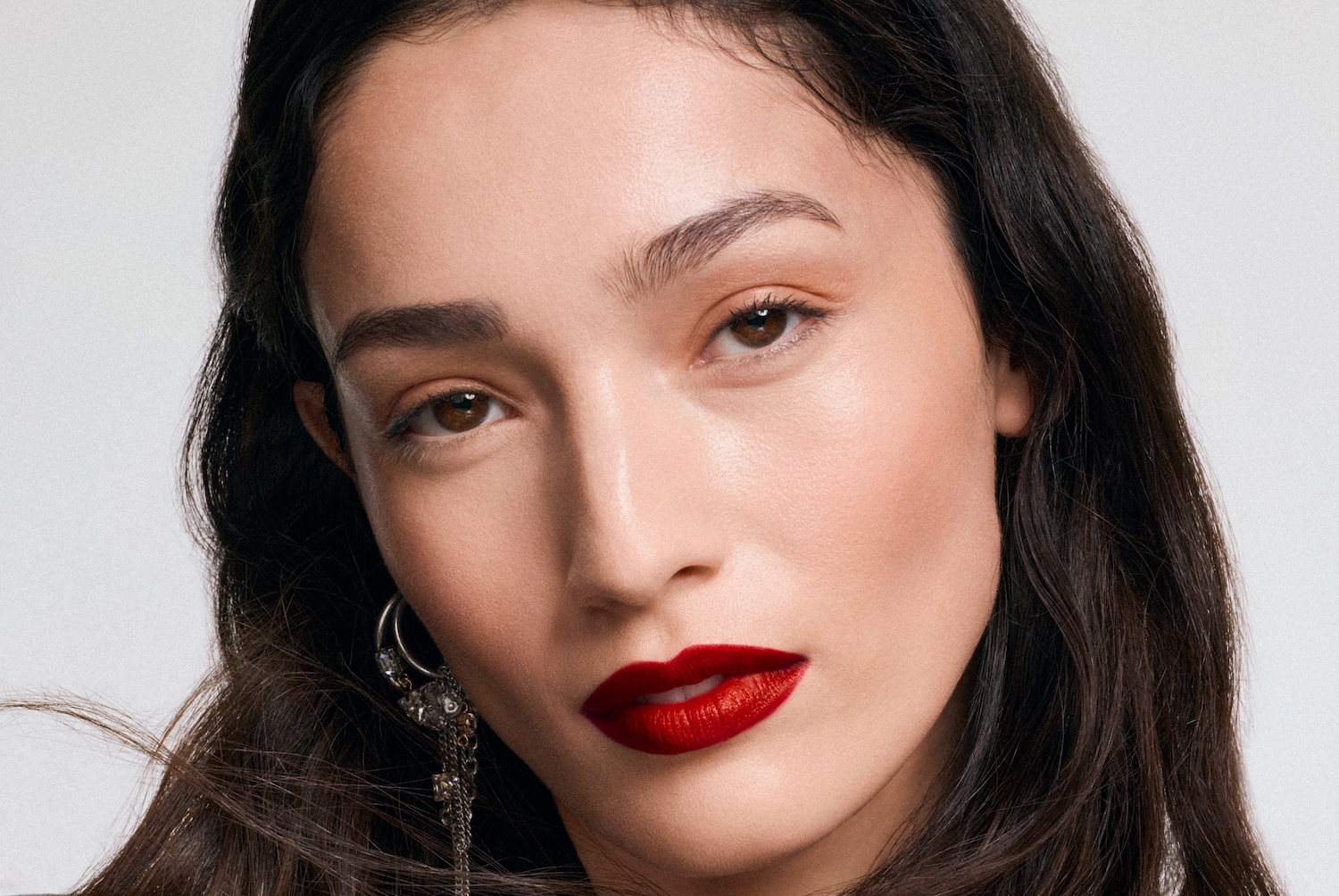 Nahaufnahme einer Frau mit braunen Augen, dunklem, gewelltem Haar und einem roten Lippenstift, die einen silbernen Ohrring mit Strasssteinen und Ketten trägt.