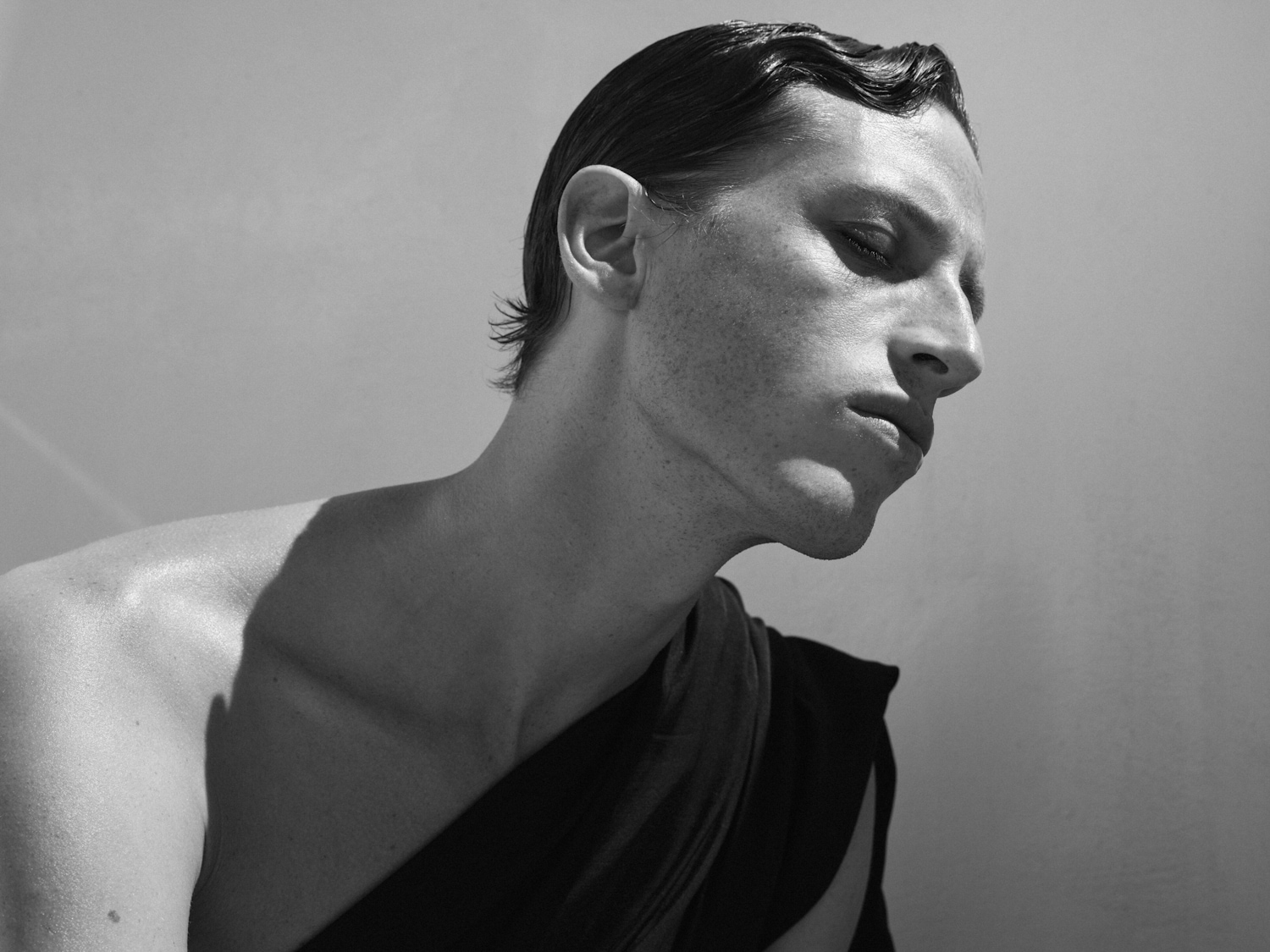 En una imagen en blanco y negro, un hombre de piel clara, con el pelo peinado hacia atrás y vestido con una túnica oscura, mira hacia abajo con los ojos cerrados.