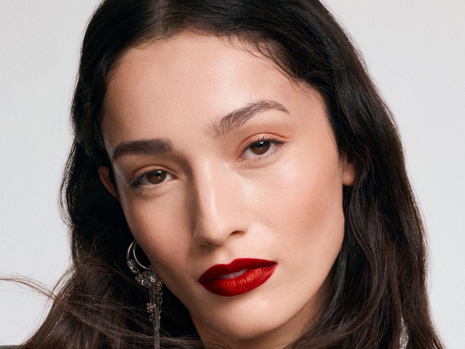 Nahaufnahme einer Frau mit dunklen, gewellten Haaren, braunen Augen und einem auffälligen roten Lippenstift, der ihren Look hervorhebt.