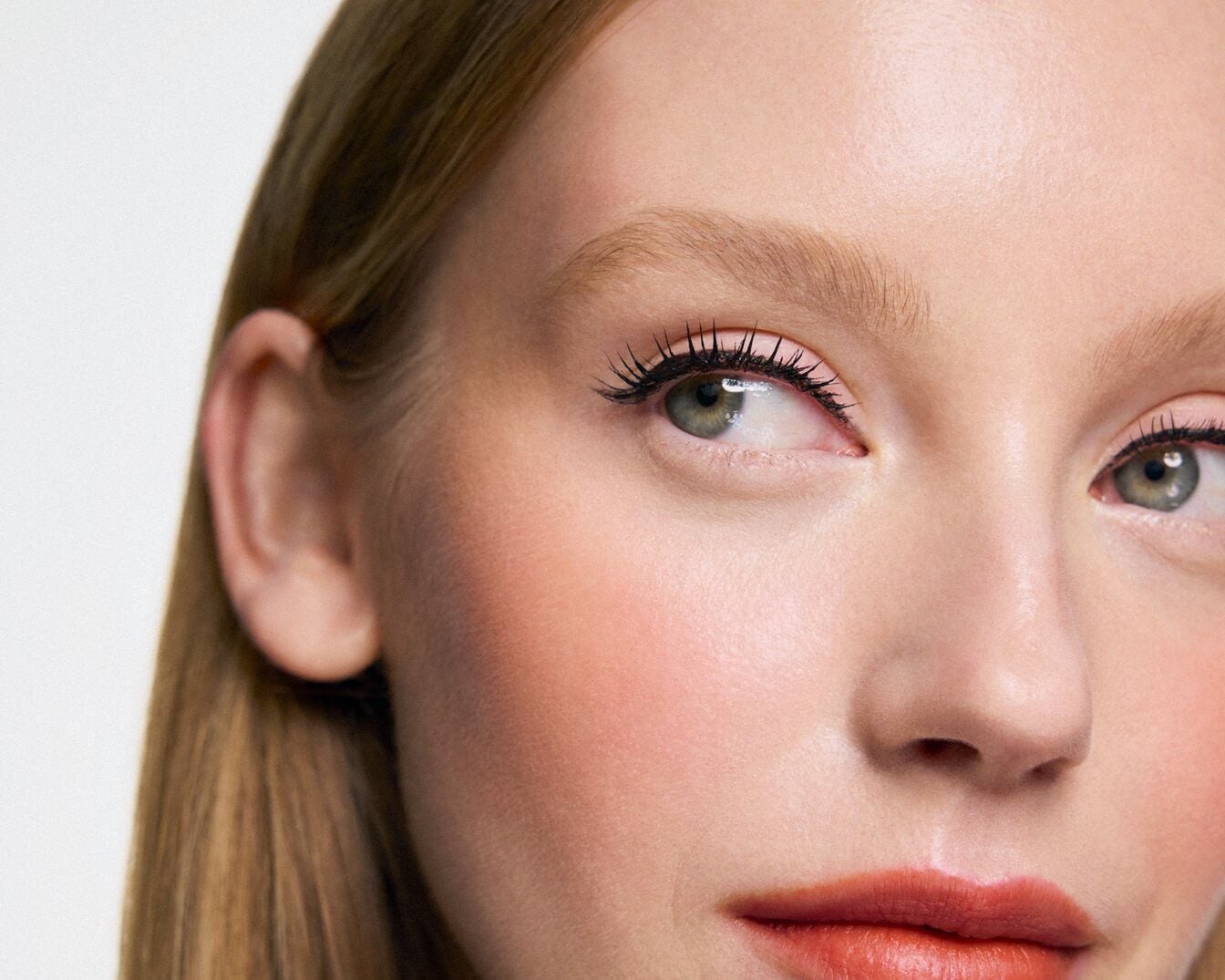 Nahaufnahme eines hellhäutigen Models mit grünen Augen, betont durch schwarze Wimperntusche, und einem Hauch von Rouge auf den Wangen, abgerundet durch einen korallfarbenen Lippenstift.