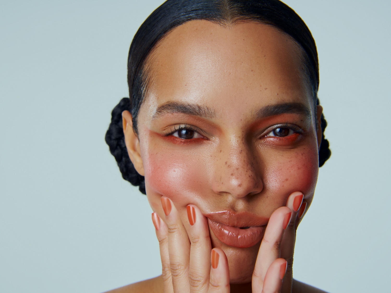 Nahaufnahme eines Models mit orangefarbenem Lidschatten und Rouge, die Hände an den Wangen, um einen frischen, strahlenden Look zu präsentieren.