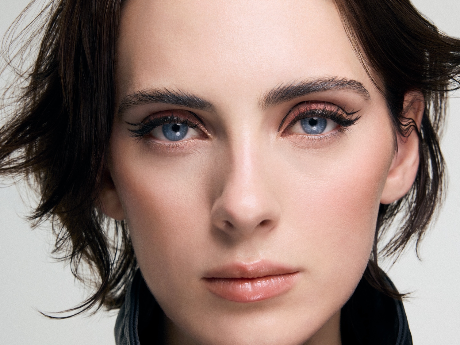 Nahaufnahme eines Frauengesichts mit hellblauen Augen, zarten rosa Wangen und glänzenden Lippen, umrahmt von dunklem, gewelltem Haar.